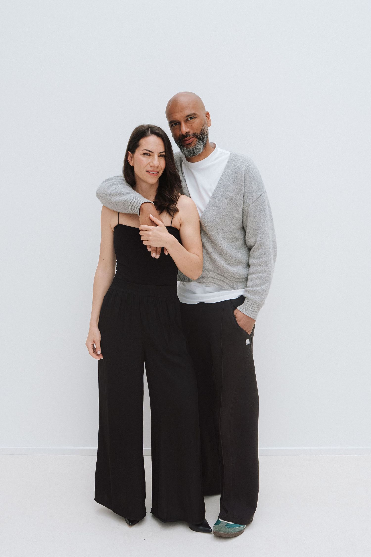 A man and woman pose together in matching black wide-leg pants against a white background.