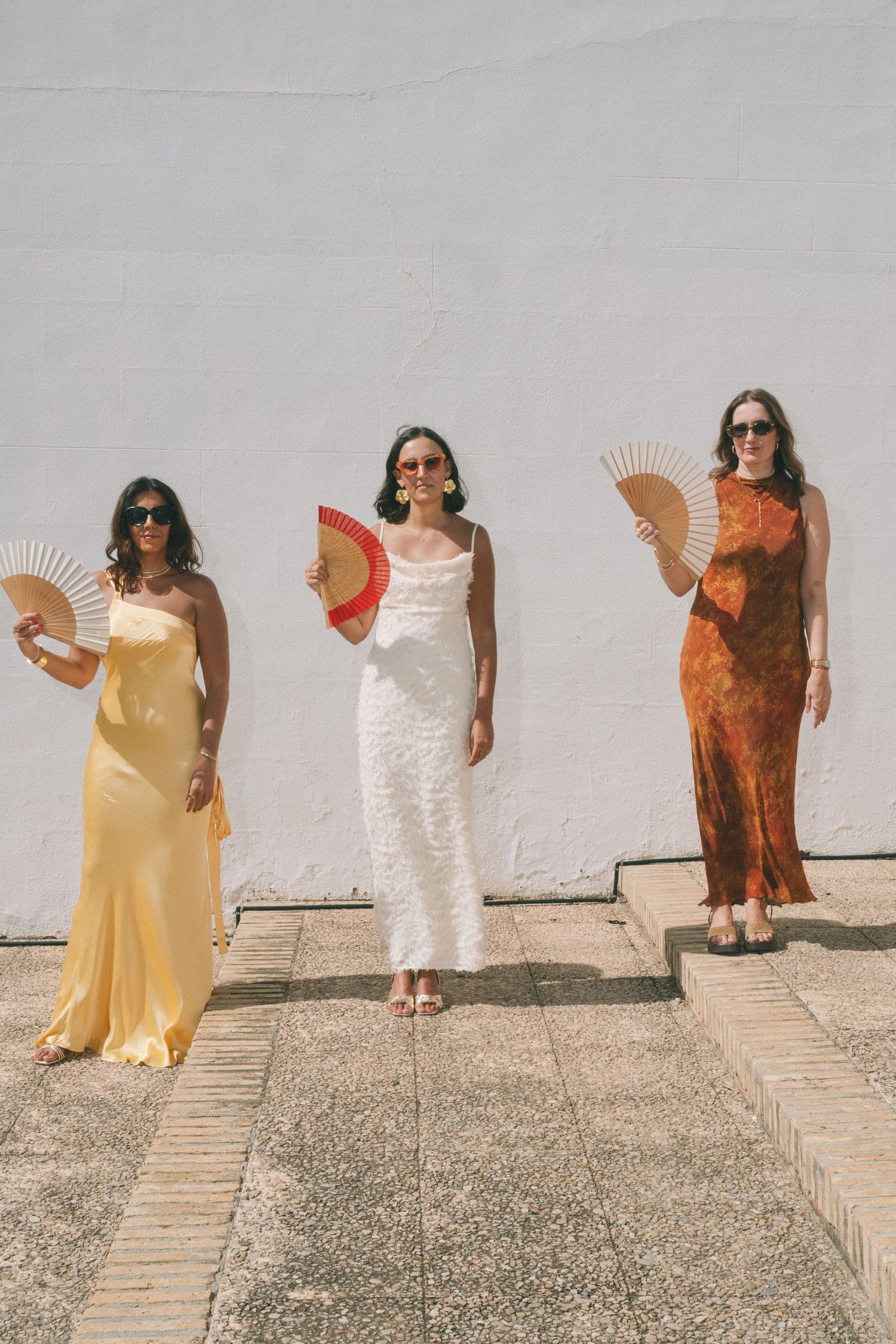 Row of guests in colorful dresses holding decorative fans while posing against a white wall.