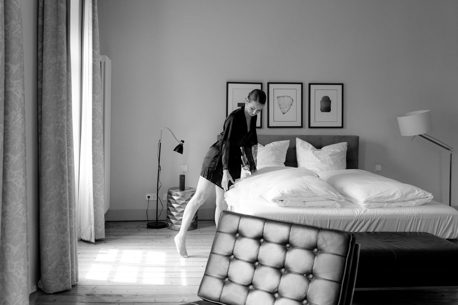 Modern minimalist bedroom interior with white bedding, wooden floors, and natural light streaming through windows.