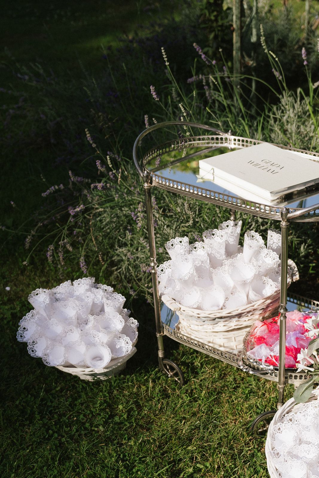 Elegant outdoor wedding display with white lace favors on a gold tiered cart, guest book, and floral arrangements on green grass.