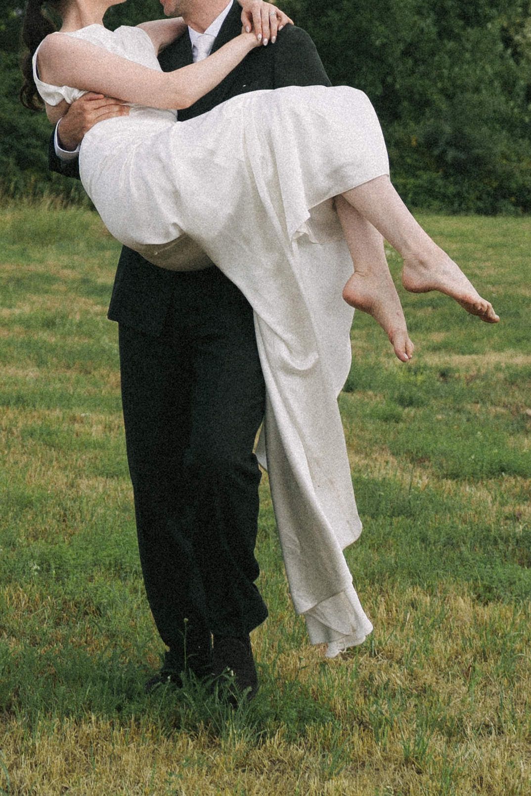 Groom in black suit carrying bride in white wedding dress outdoors on green grass field.