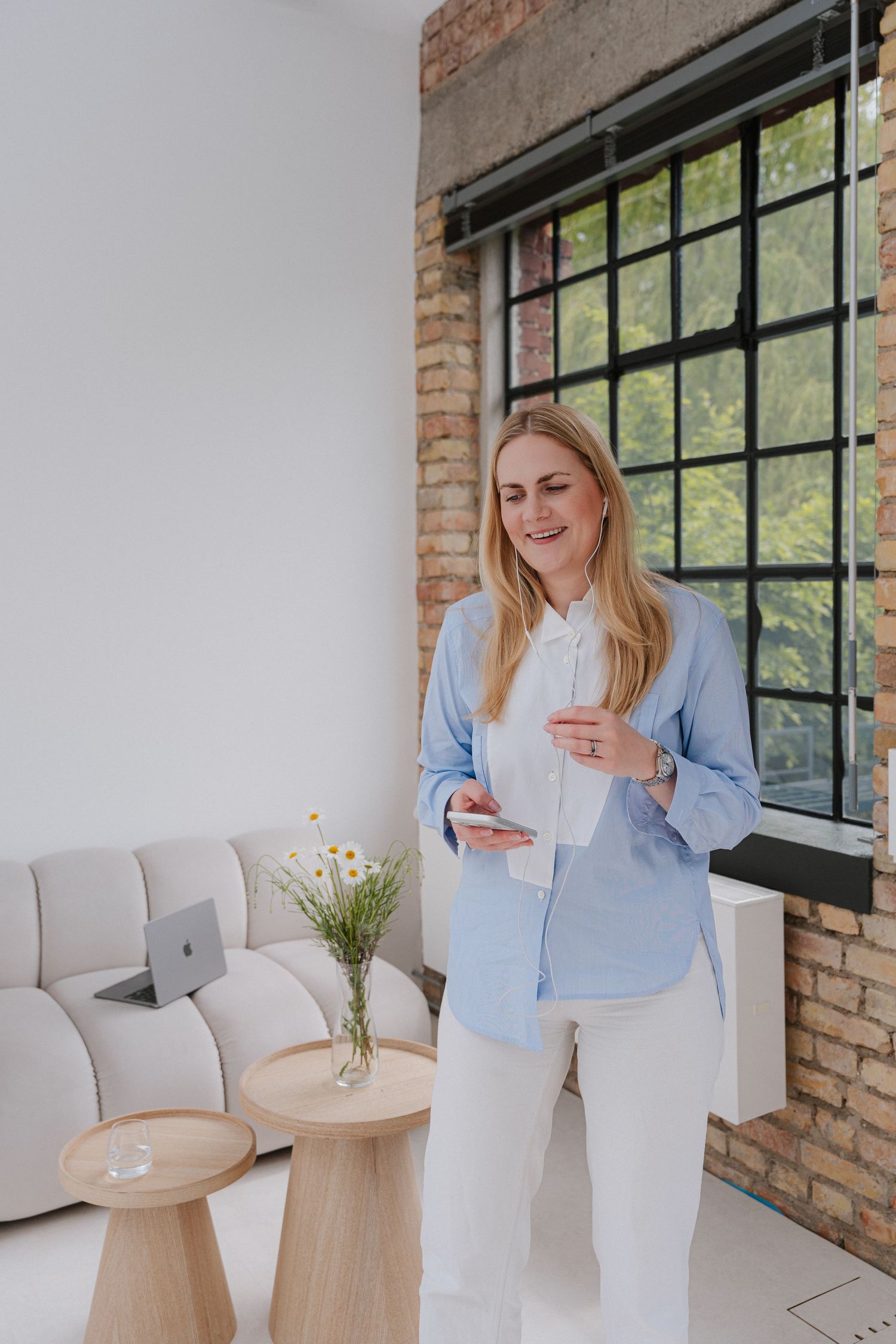 Smiling blonde woman in blue shirt and white pants using smartphone in modern loft office with brick wall and large window.