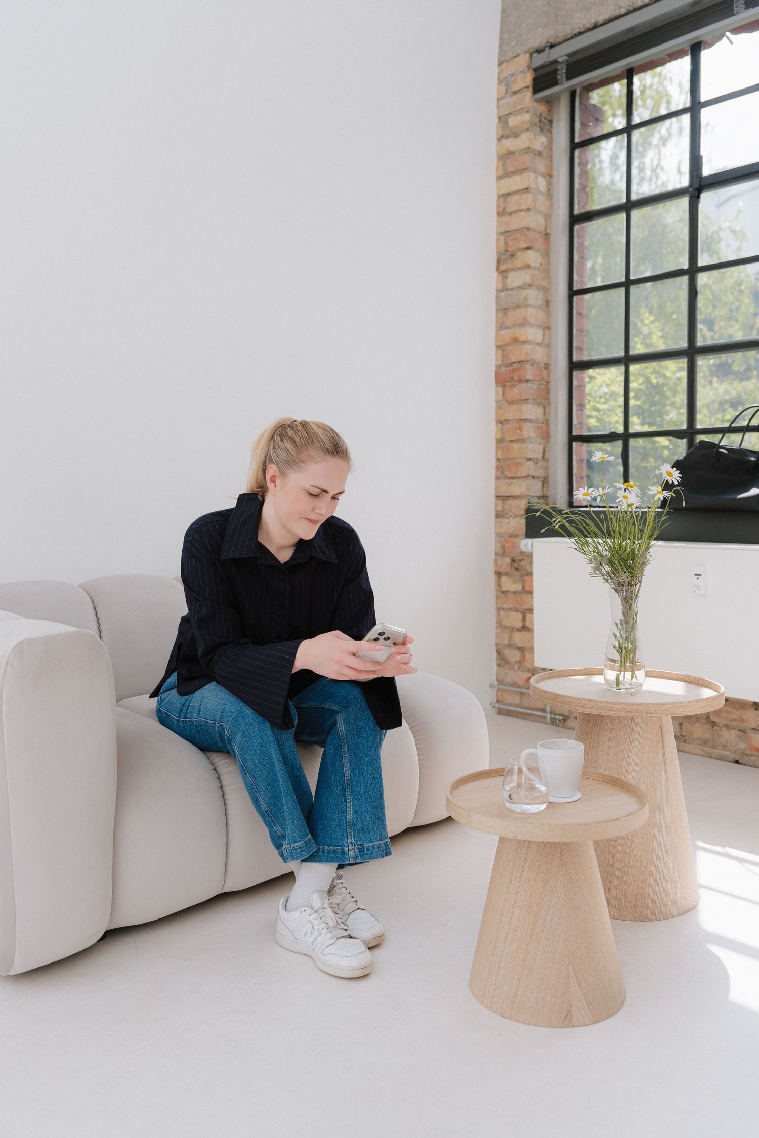 Young blonde woman in black shirt and jeans sitting on beige sofa using smartphone near wooden side tables.