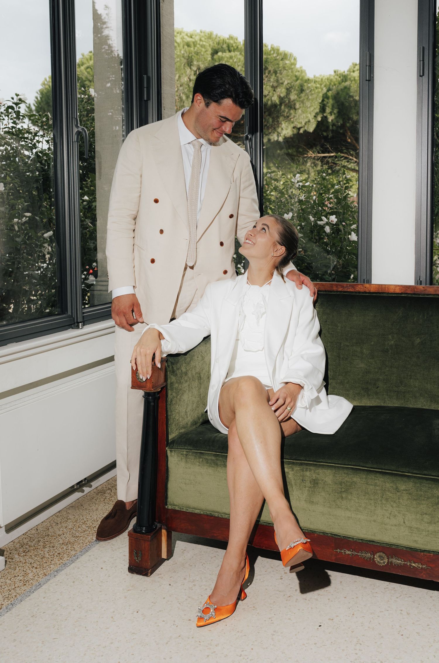 Couple in white formal attire posing playfully in sunlit room with windows.