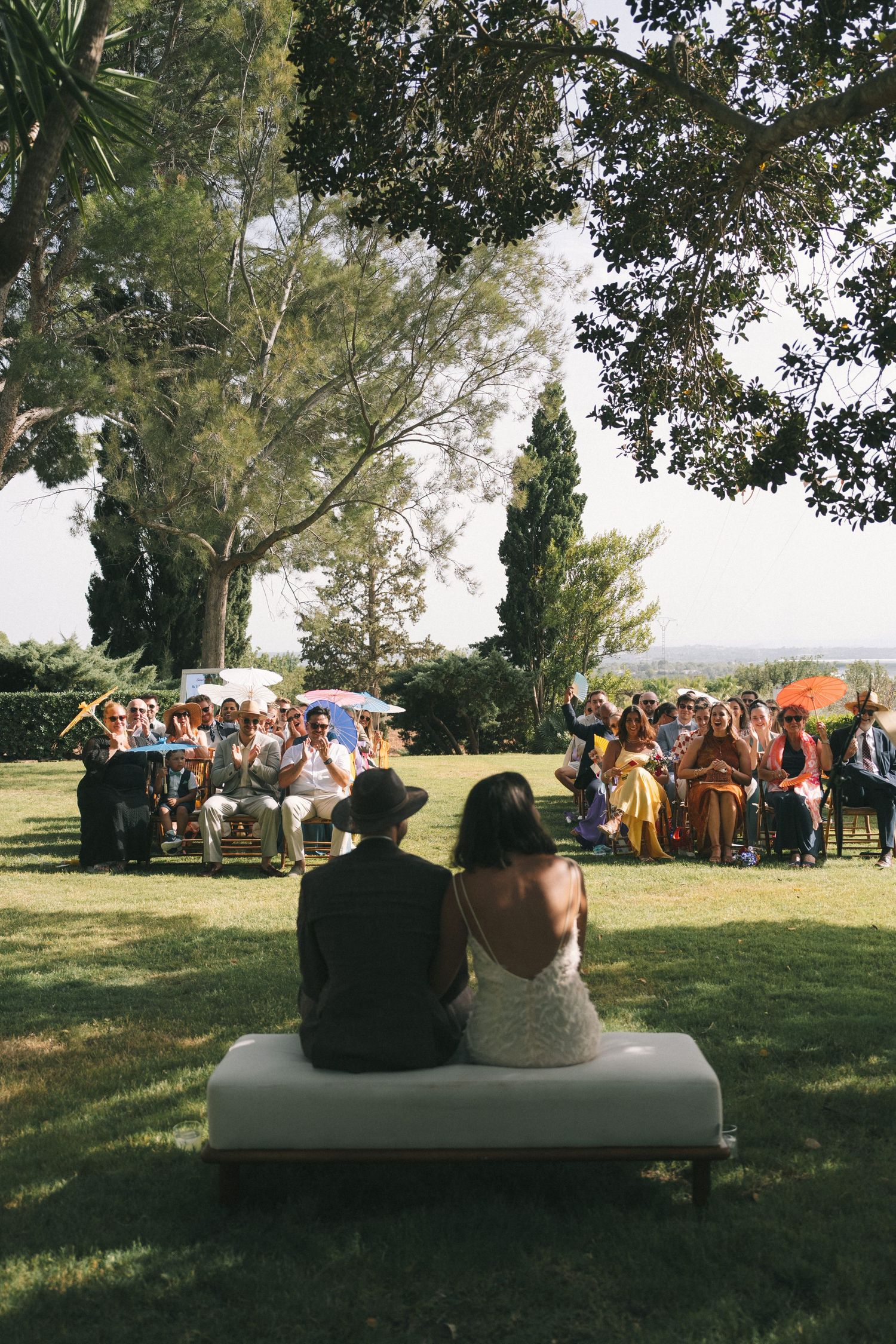 Couple sitting on bench overlooking outdoor wedding ceremony with guests in background.