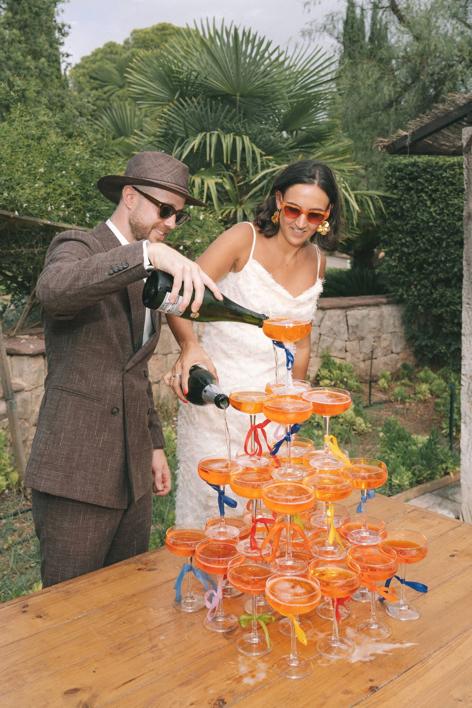 Couple pouring champagne into tower of orange cocktail glasses at outdoor celebration.
