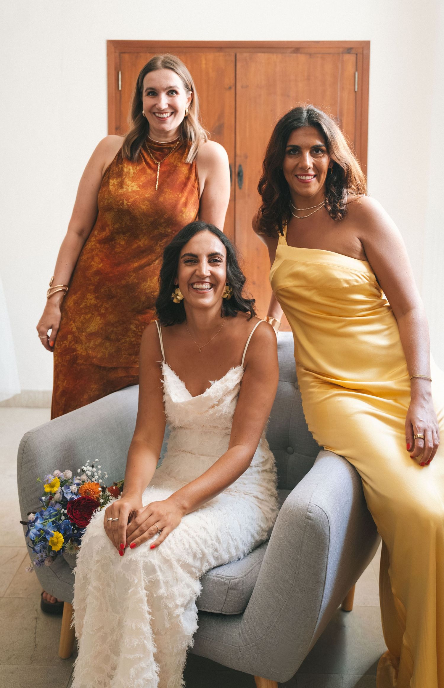 Wedding party members relax on a grey couch wearing yellow and brown dresses.