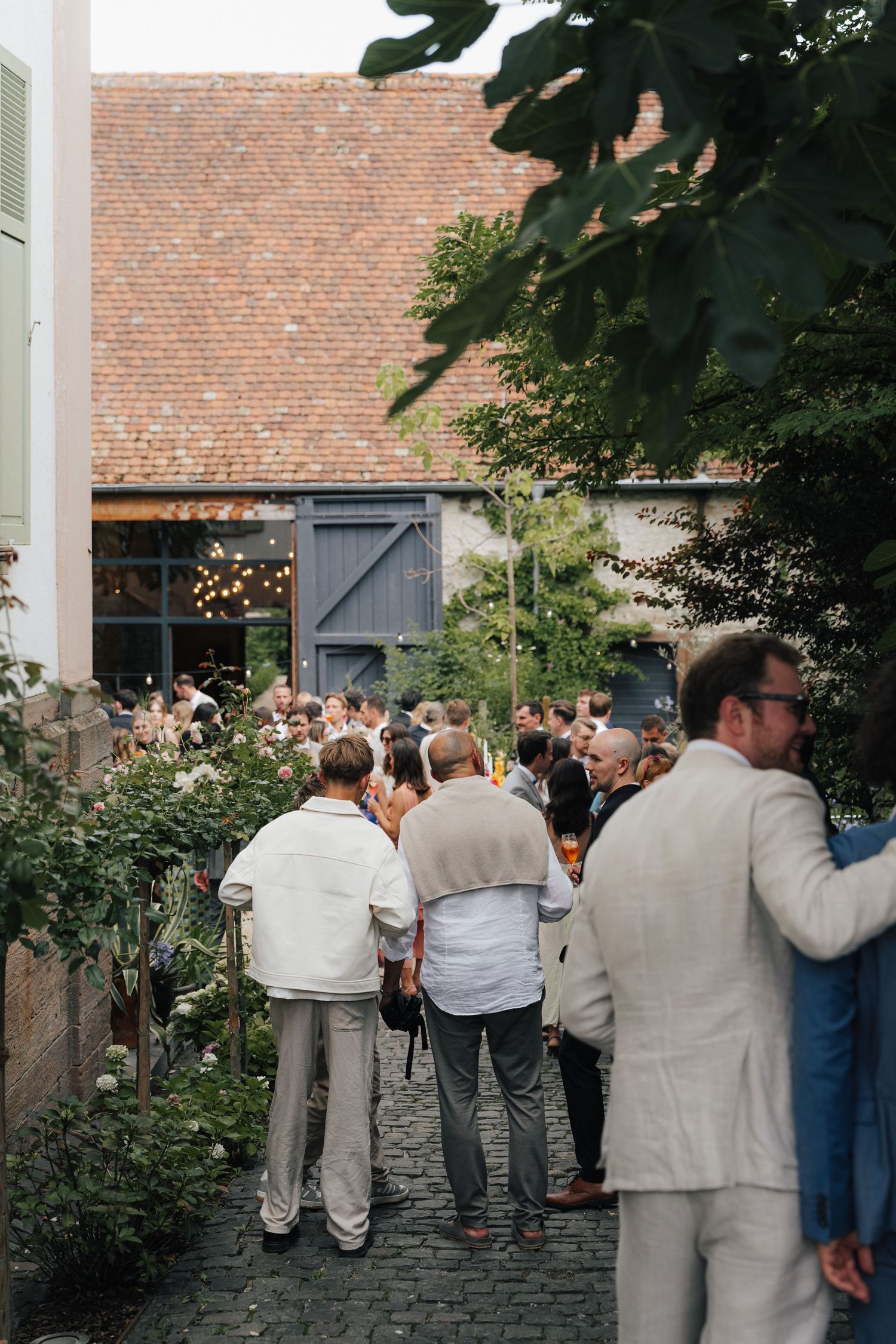 Wedding guests gather outside a rustic brick building with string lights and garden decor.