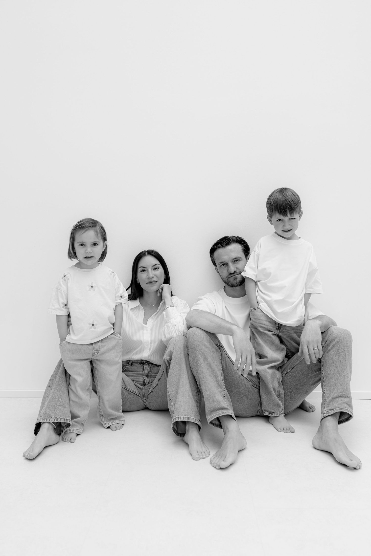 Black and white family portrait of parents and two children sitting together on white background in casual white shirts and jeans.