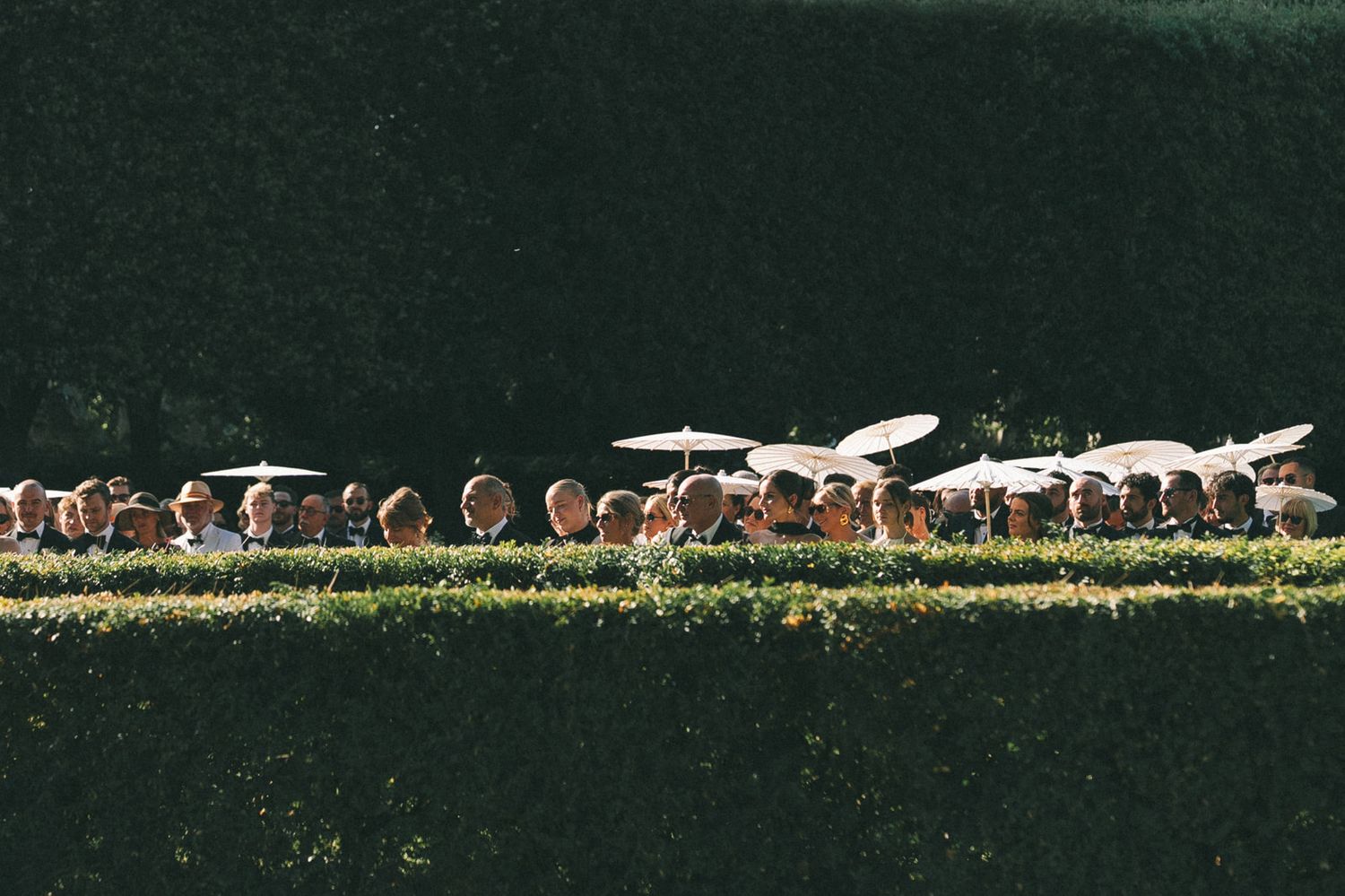 Aerial view of wedding guests seated outdoors under white parasols along a hedge-lined garden aisle.