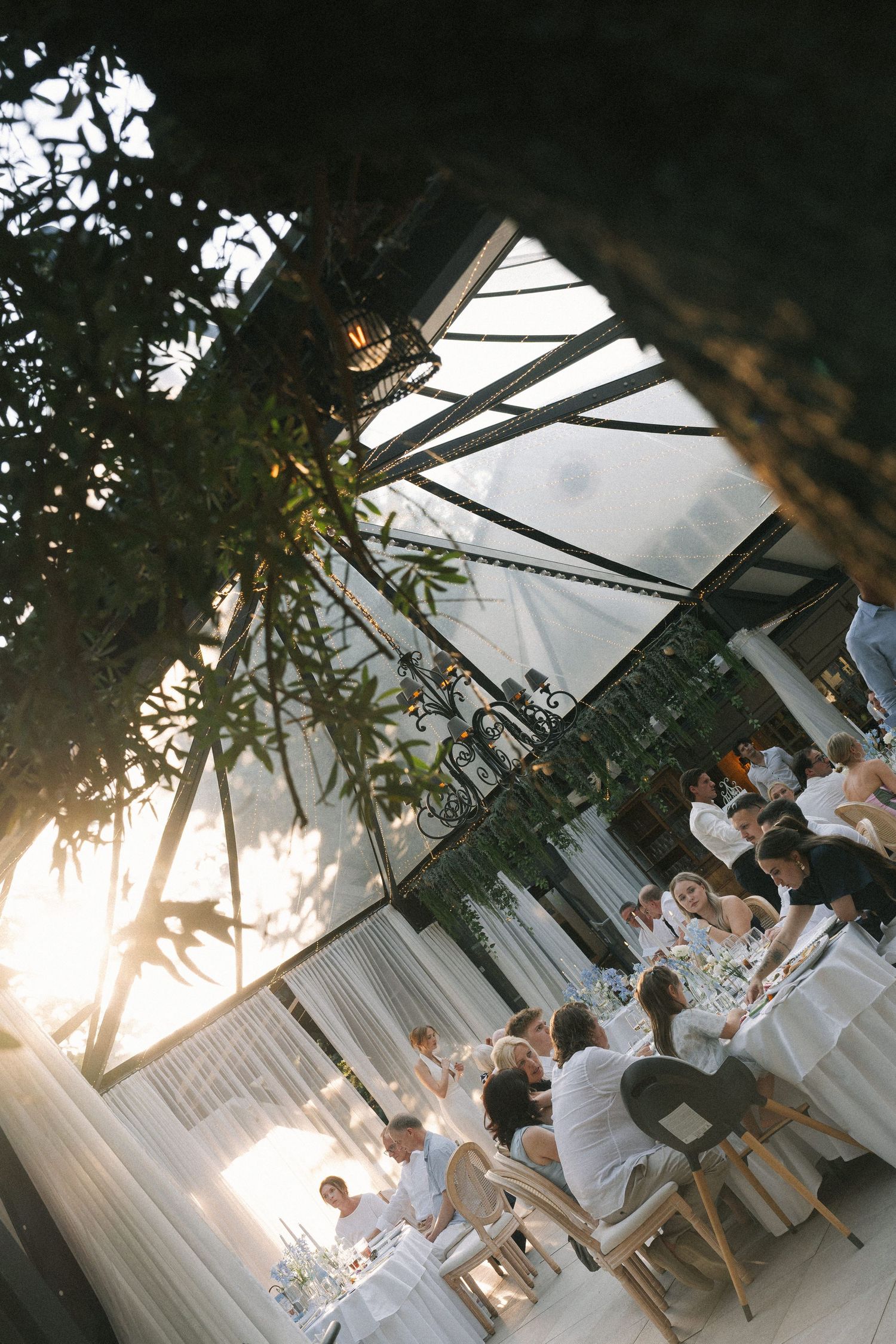 Elegant outdoor wedding reception under a glass greenhouse with guests in white at golden hour.