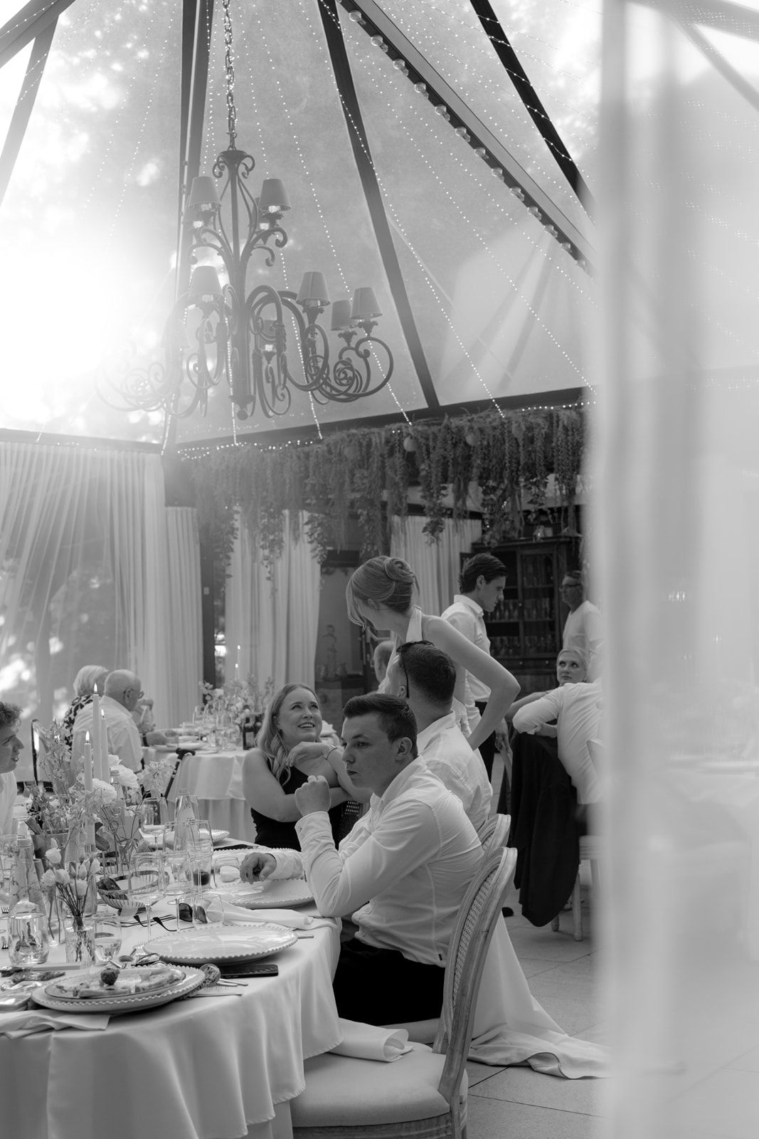 Black and white photo of elegantly dressed guests dining under a decorated tent with chandelier at a formal event.
