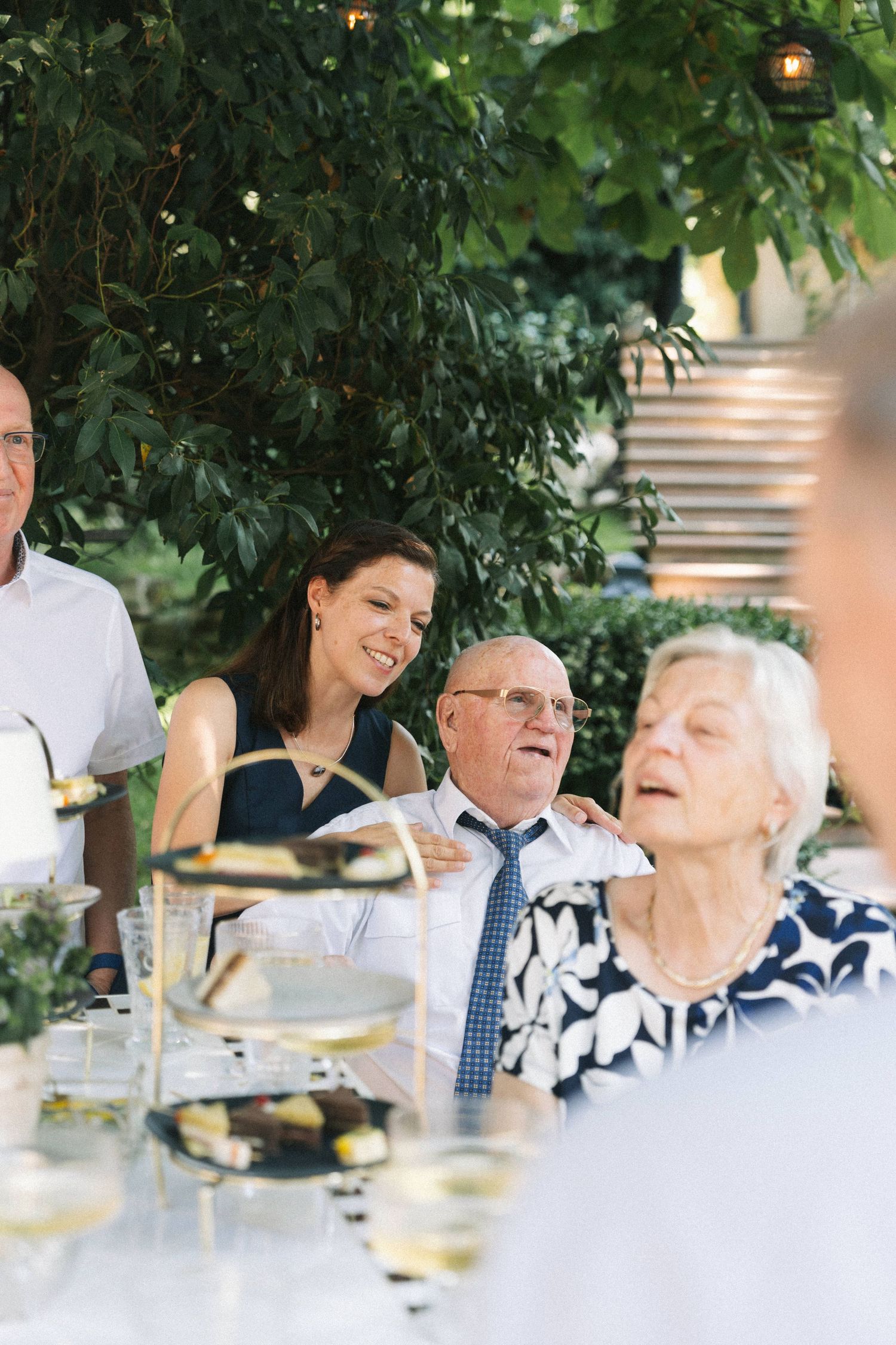 Guests smiling and laughing at an outdoor wedding reception dinner table surrounded by lush green trees.