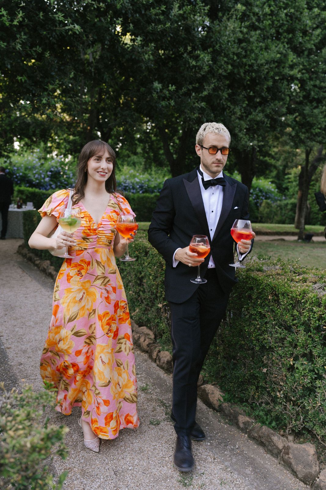 Elegant couple in formal attire holding cocktails in a lush garden, woman in floral dress, man in black tuxedo.