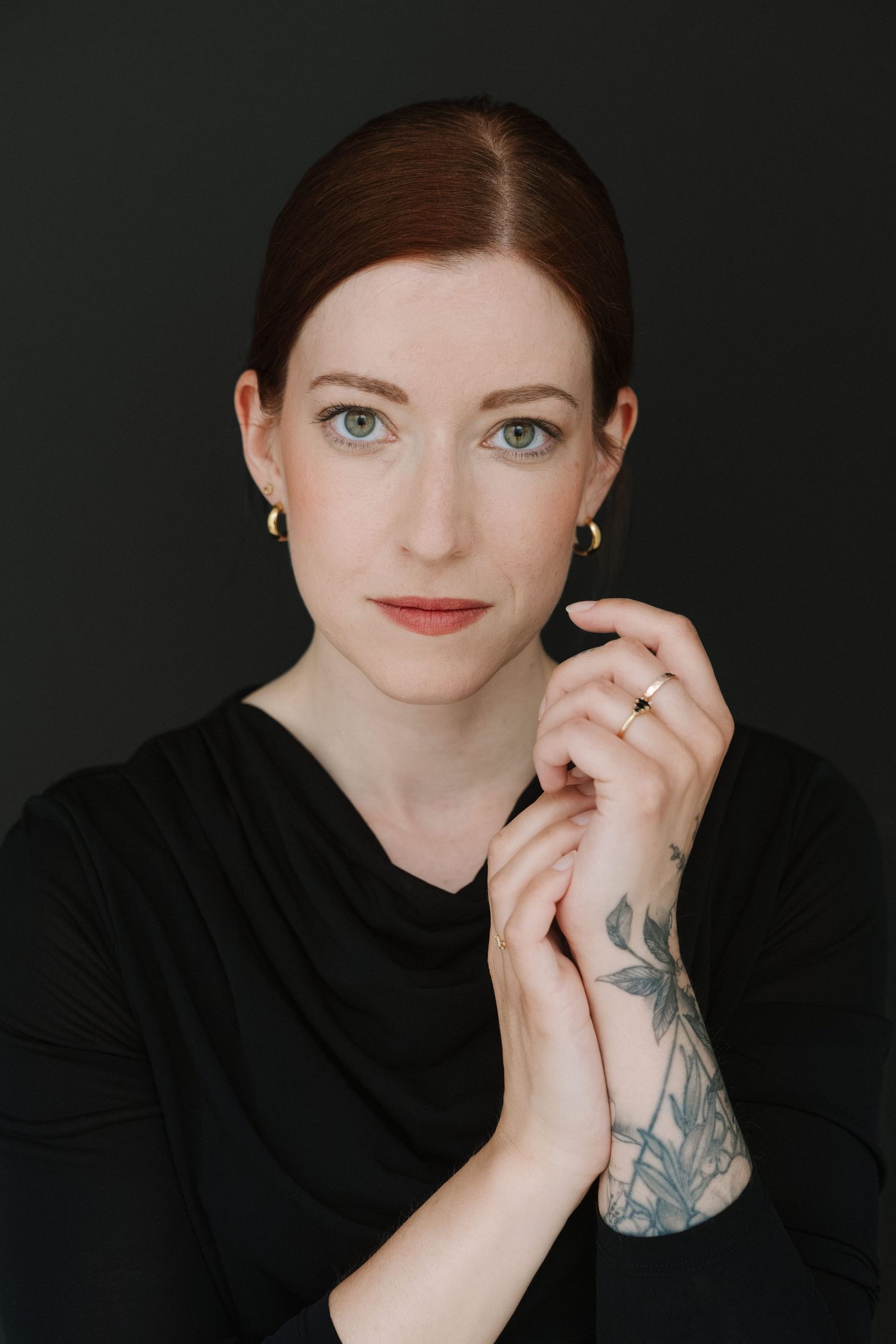 Woman with red hair, green eyes, black top, gold earrings, and floral forearm tattoo poses against dark background.