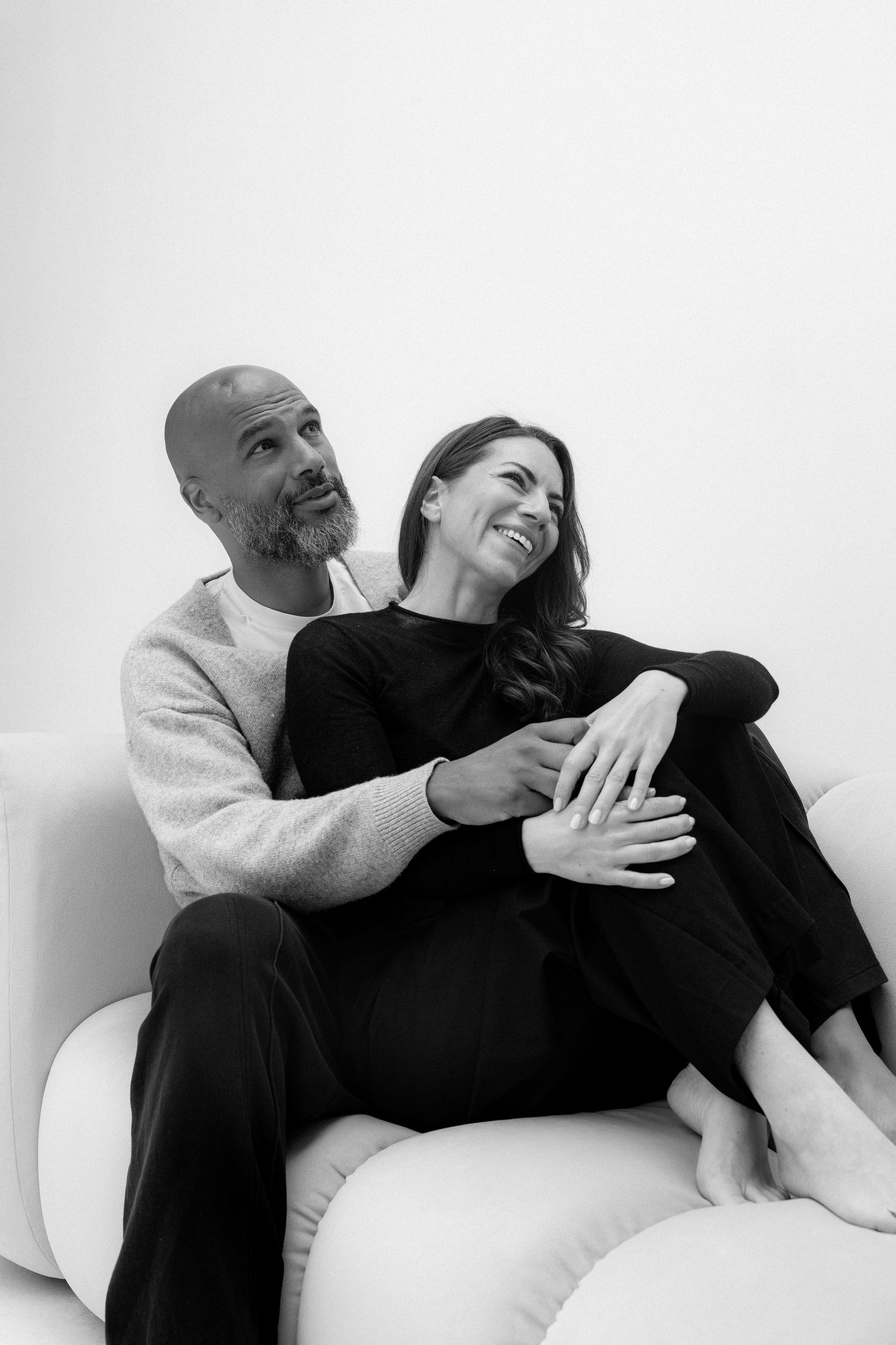 Black and white photo of a smiling couple sitting together on a white sofa, the man embracing the woman from behind.