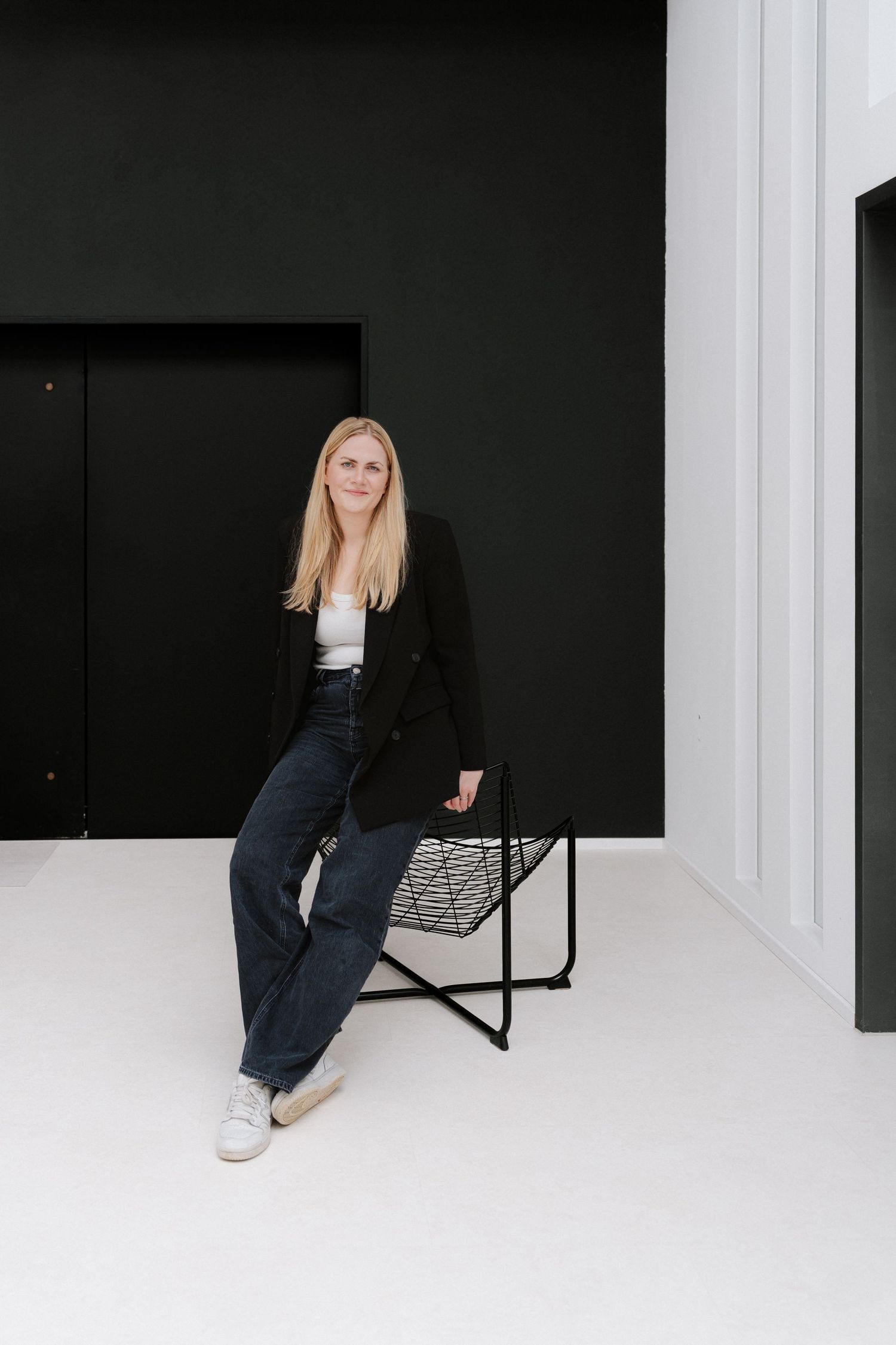 Blonde woman in black blazer and jeans sitting on modern wire chair against dark wall in minimalist white studio.