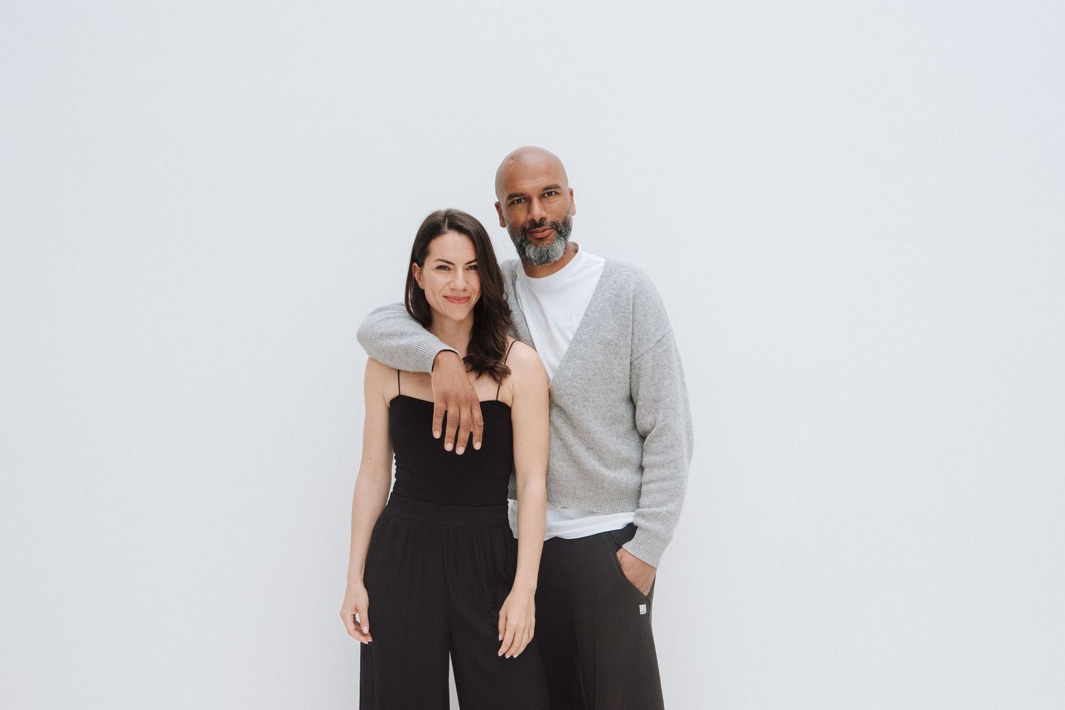A man and woman pose together smiling against a white background, dressed in casual black and gray outfits.