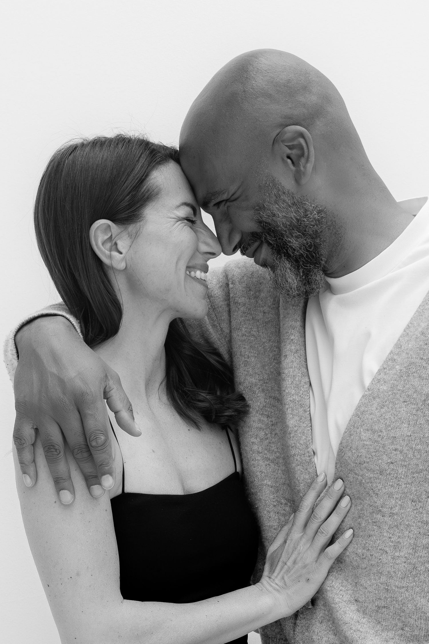 Black and white photo of a joyful interracial couple embracing, foreheads touching, smiling warmly against a white background.