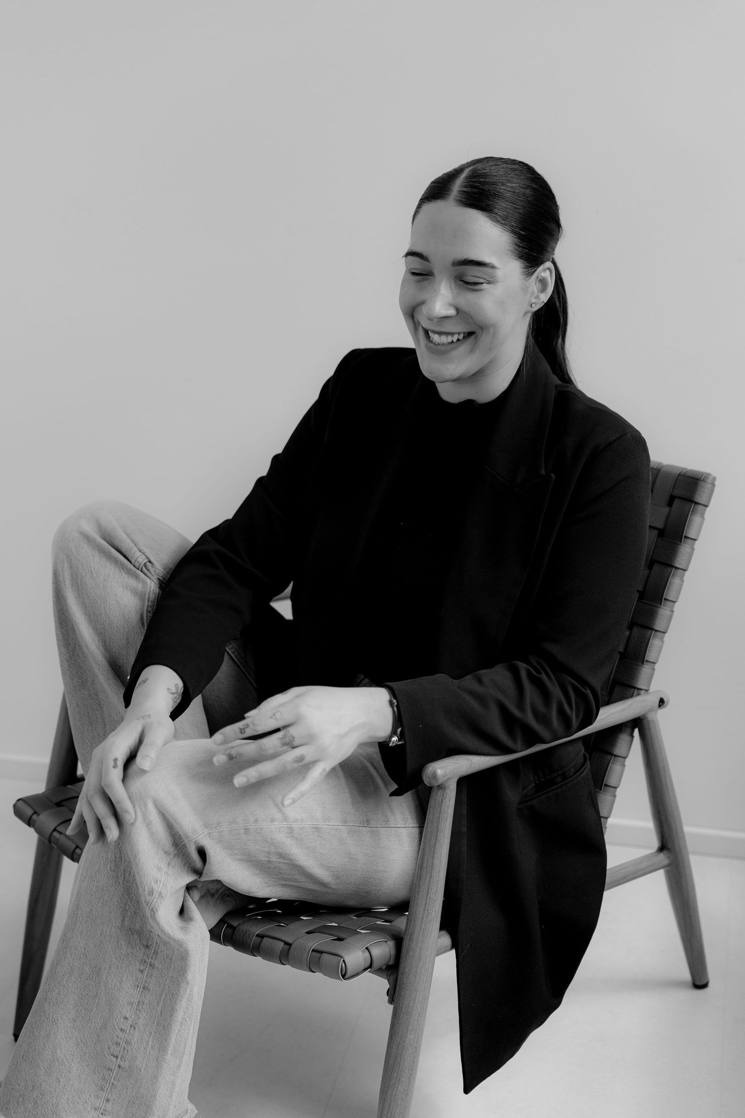 Black and white photo of a smiling woman in a black blazer sitting relaxed in a wooden chair.