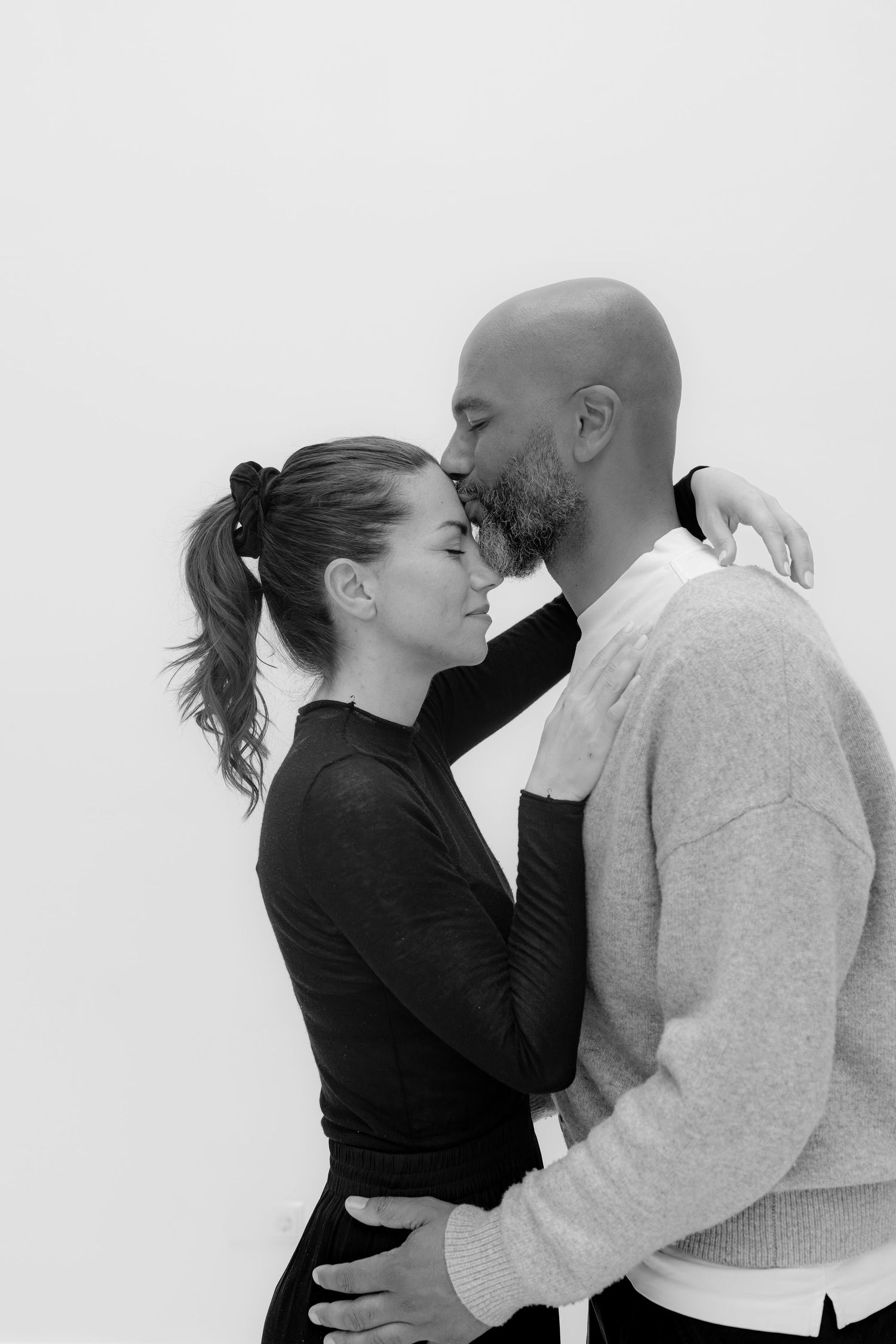 Black and white photo of a couple embracing intimately, man kissing woman's forehead against white background.