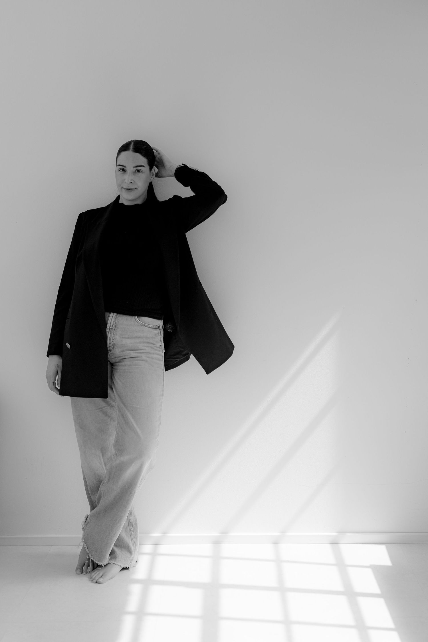 Black and white portrait of a woman in blazer and wide-leg pants leaning against a white wall in natural light.