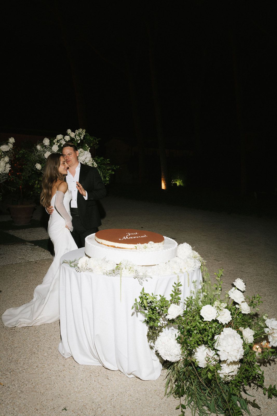 Bride and groom embrace behind wedding cake table adorned with white florals and greenery at outdoor nighttime reception.
