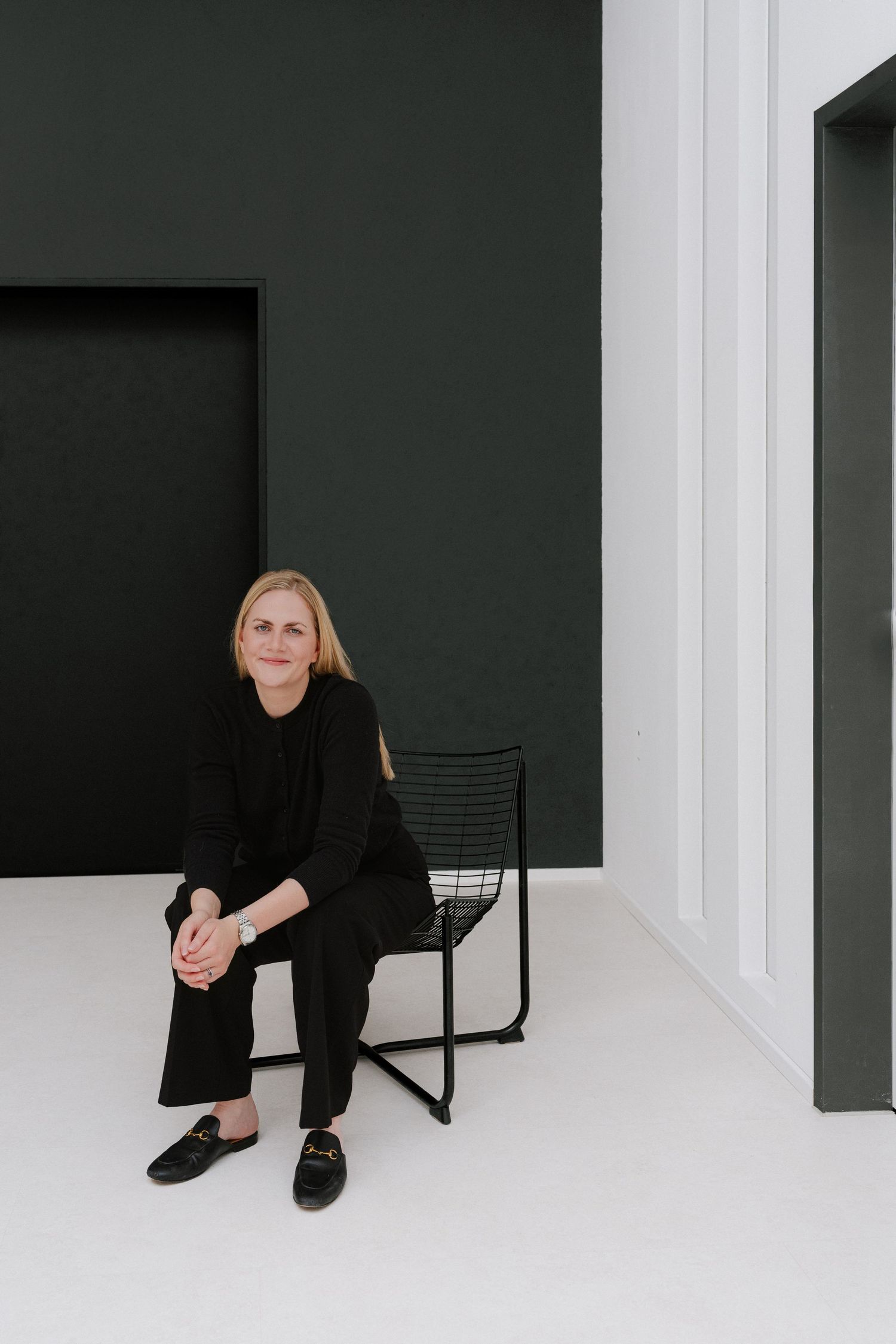 Woman in black outfit sitting on a wire chair against a dark green and white minimalist wall backdrop.
