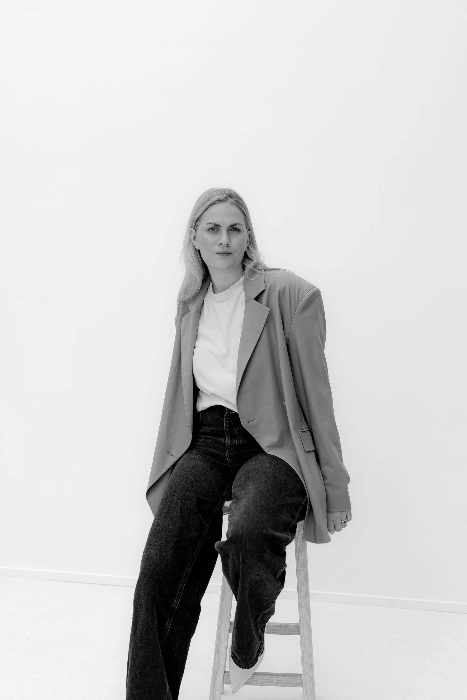 Black and white portrait of a blonde woman in blazer and jeans seated on a stool against white background.