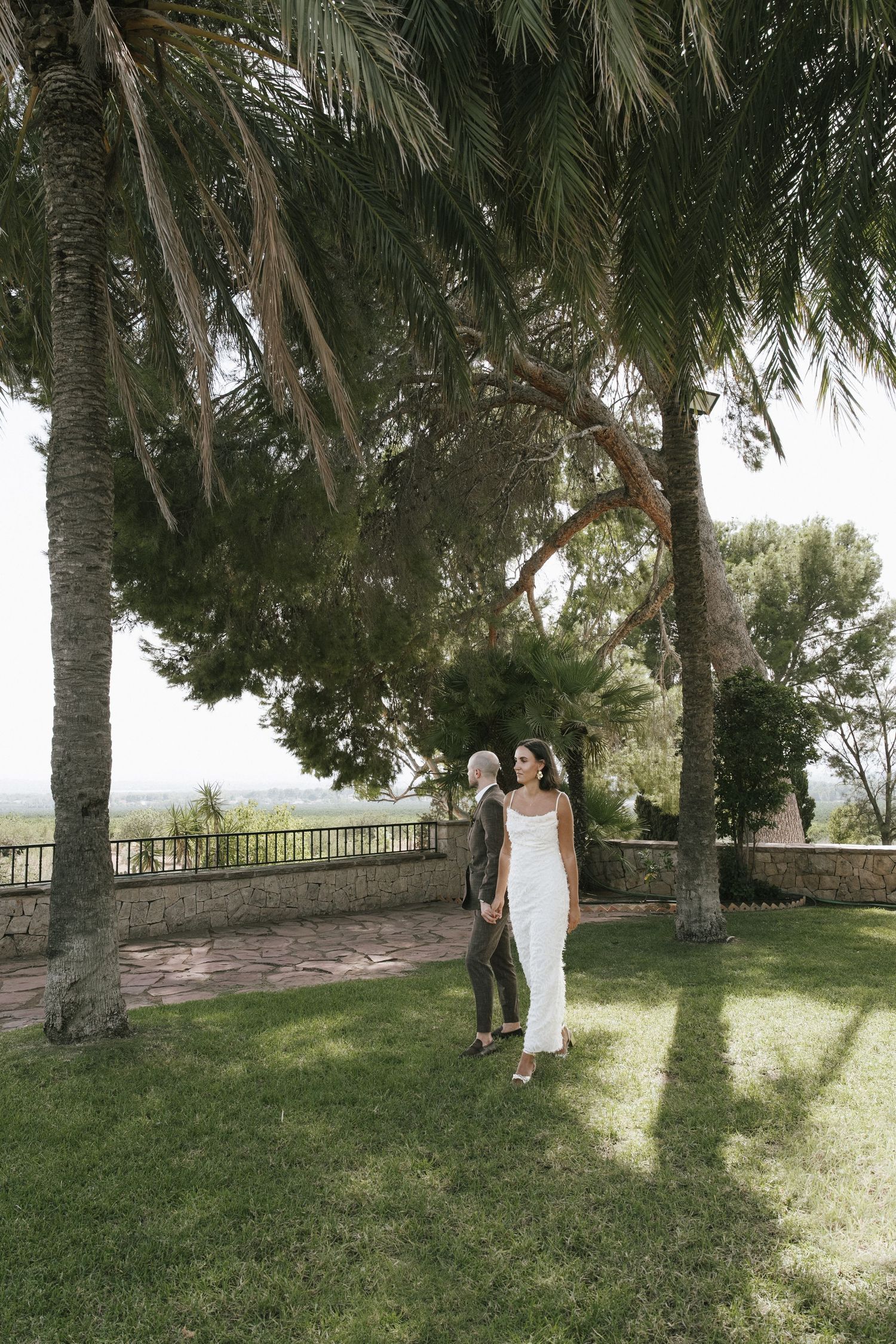 Couple walking through a lush garden with palm trees and stone pathways.