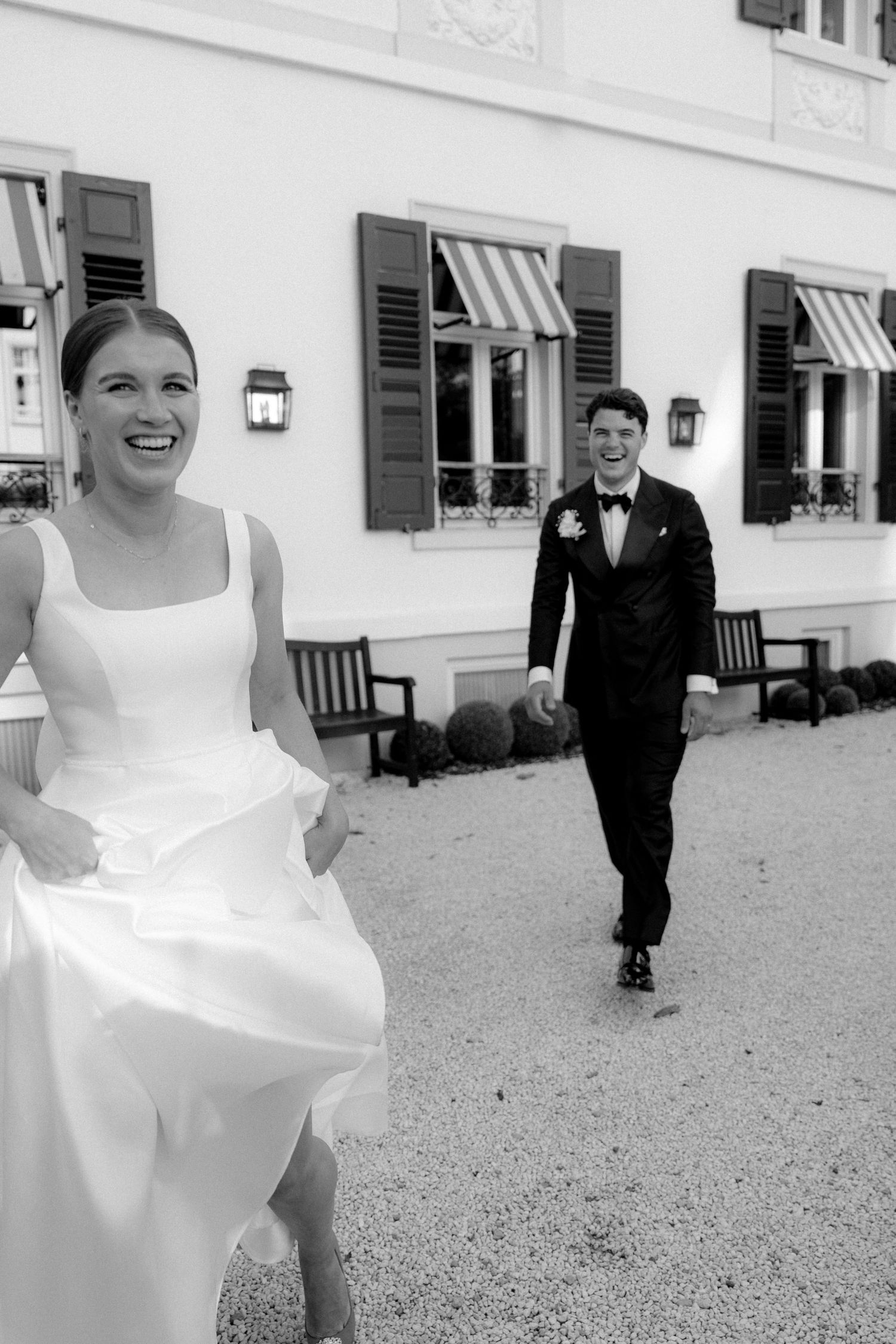 A bride in a flowing white dress twirls joyfully on a gravel courtyard.