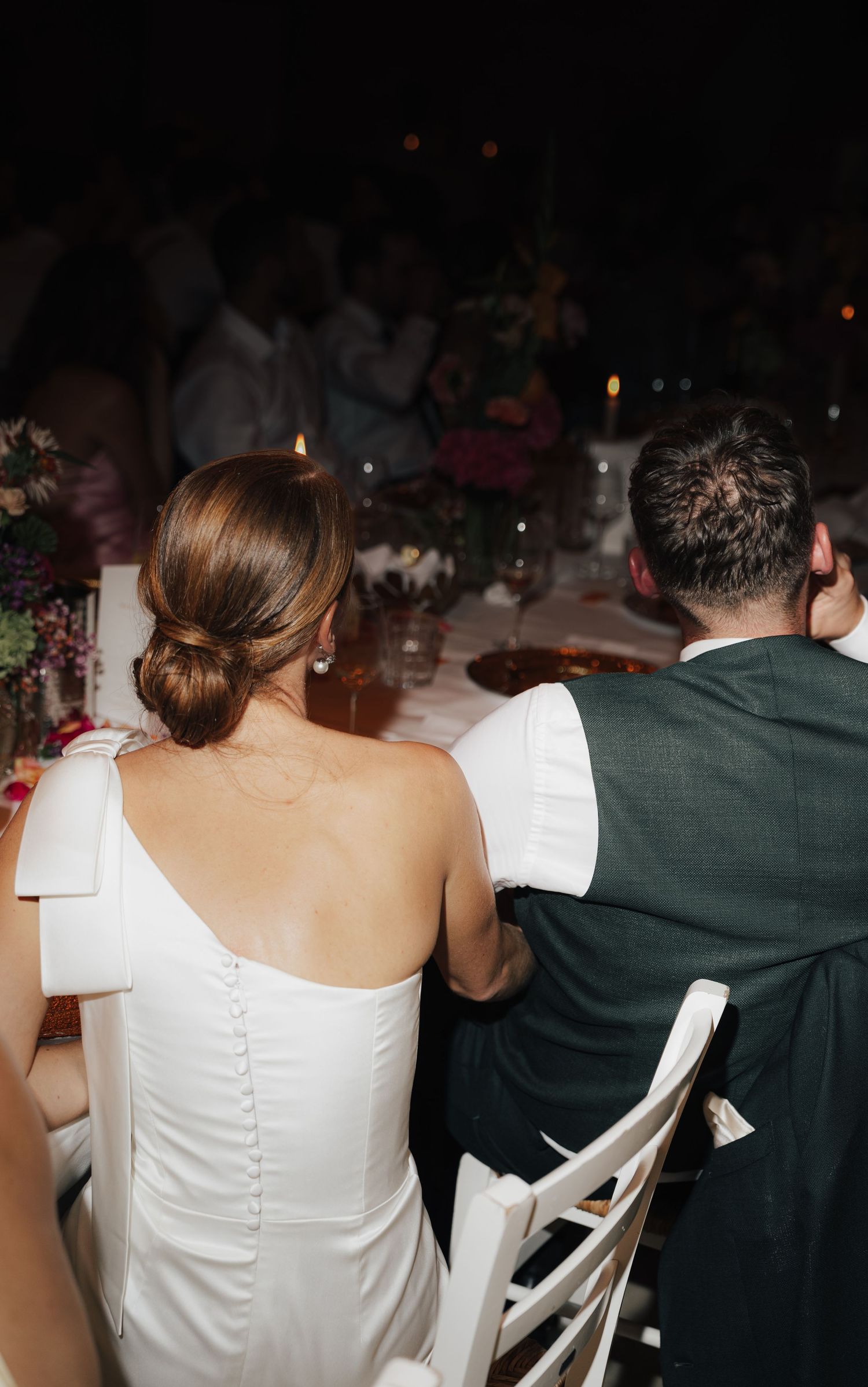 Wedding guests seated at reception tables with white chairs and dark decor visible from behind.