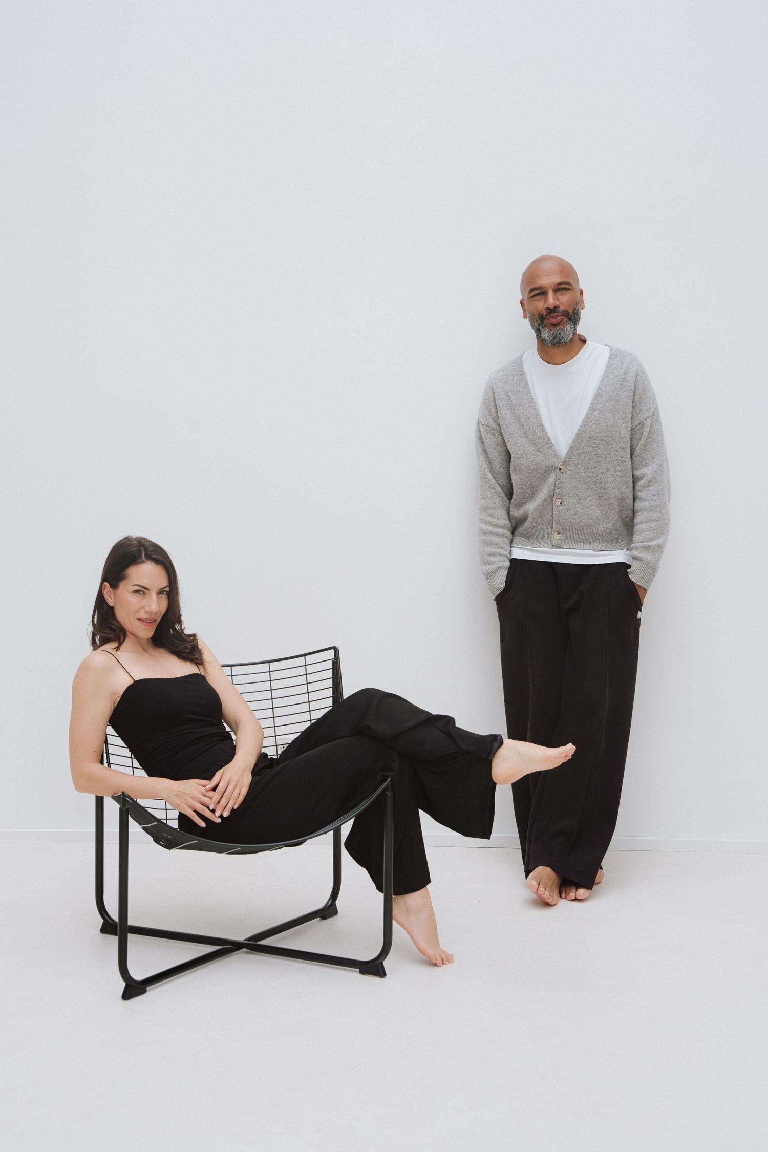 A woman in black sits in a wire chair beside a bald man in grey cardigan and black pants, both barefoot on white background.