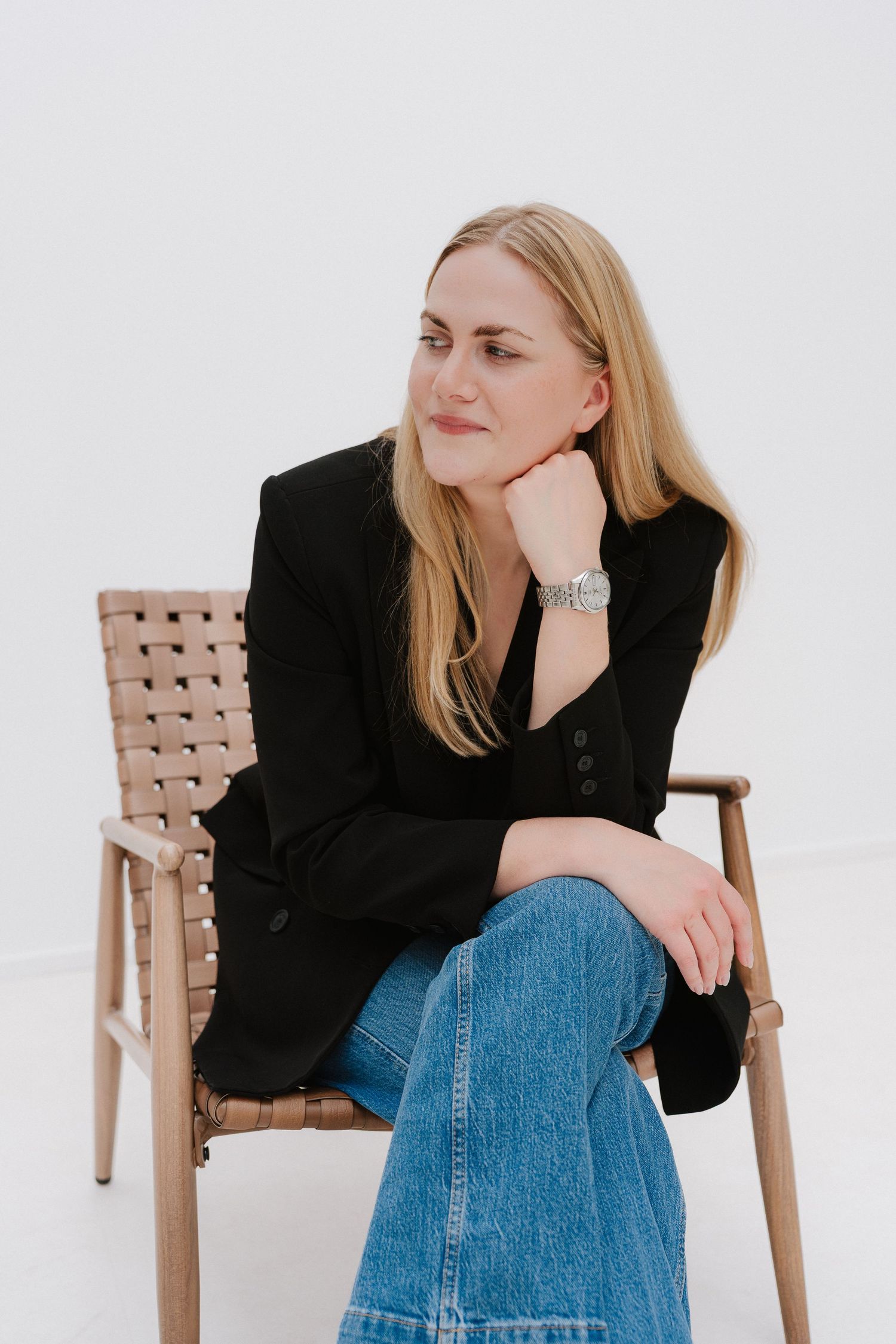 Blonde woman in black blazer and jeans sits relaxed on wooden chair, resting chin on hand, against white background.