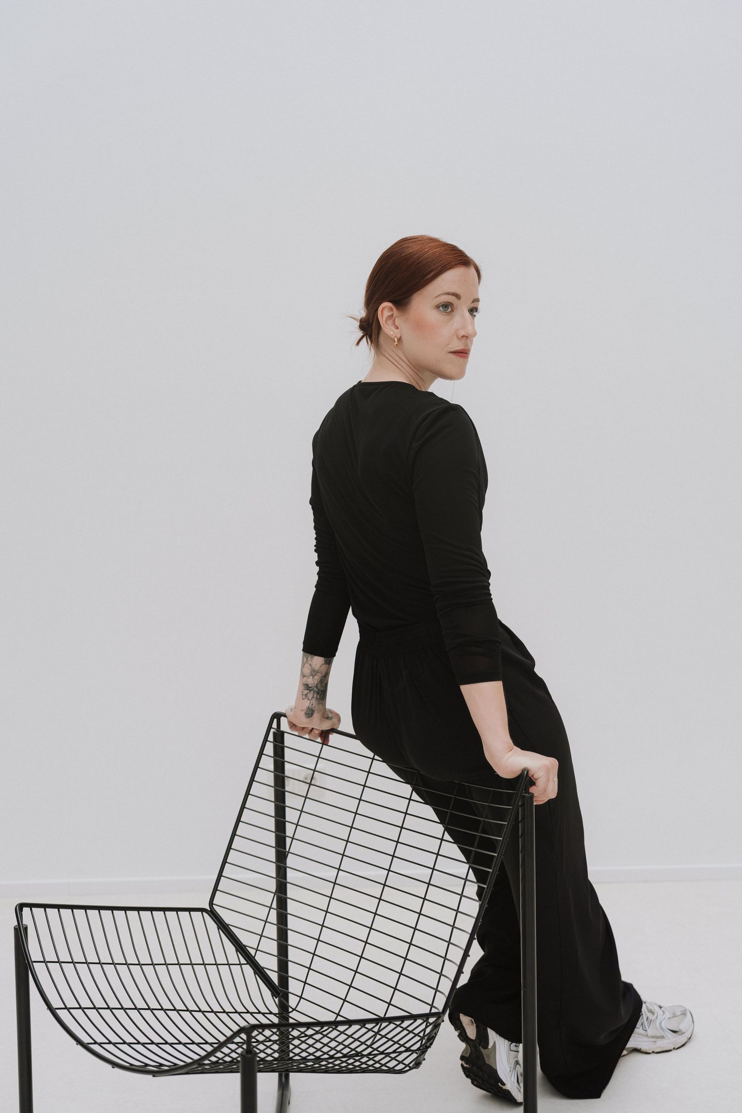 Woman in black dress with tattoos poses with wire metal chairs against white background.
