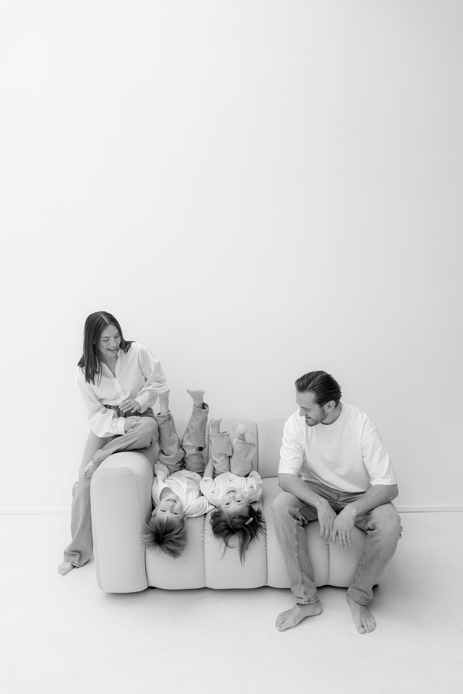 Black and white photo of a family of four on a white couch, kids lying upside down while parents watch them play.