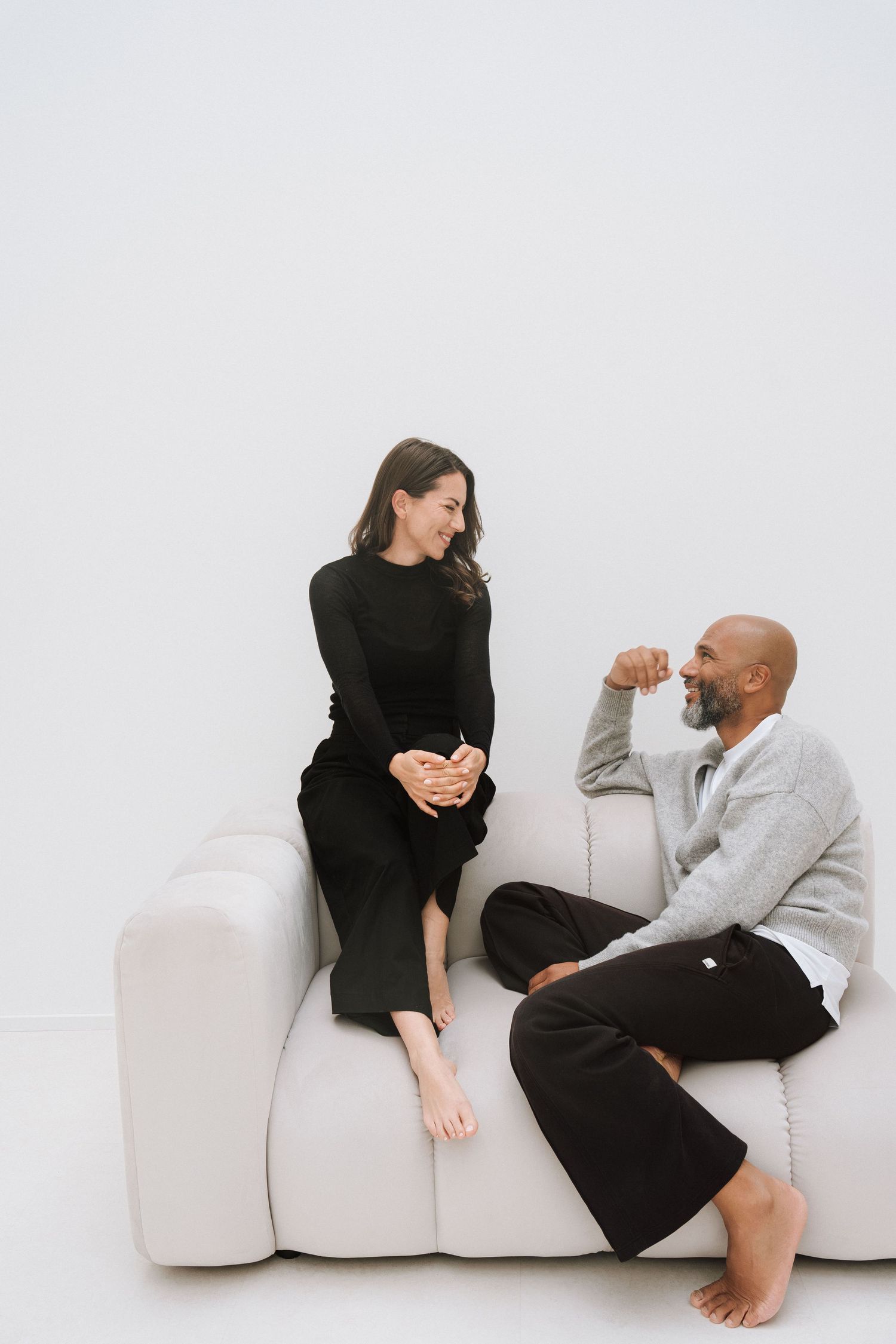A woman in black dress and man in gray sweater sit barefoot on a white sofa, smiling and conversing together.