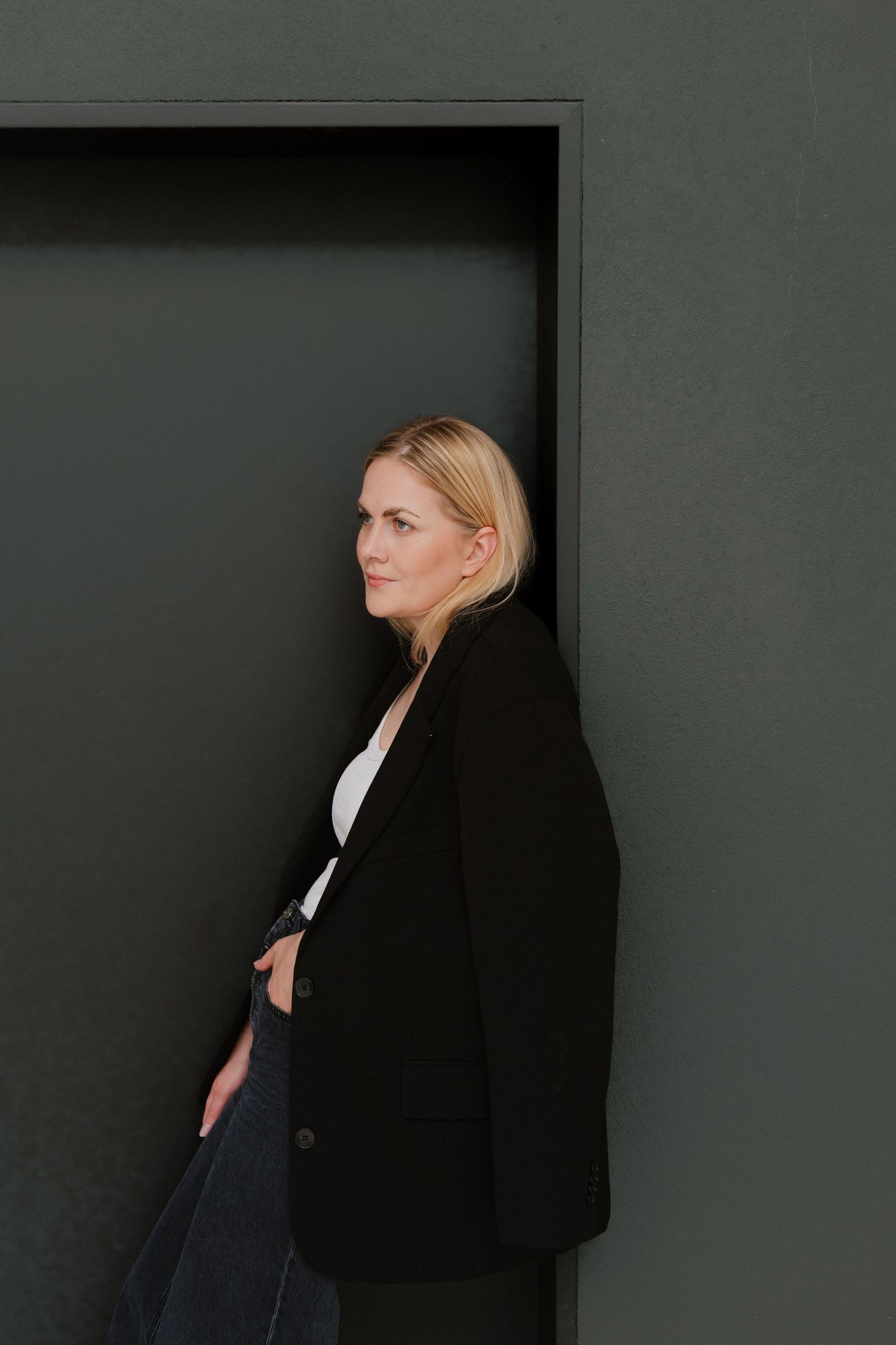 Blonde woman in black blazer leaning against dark green wall, looking sideways in a contemplative pose.