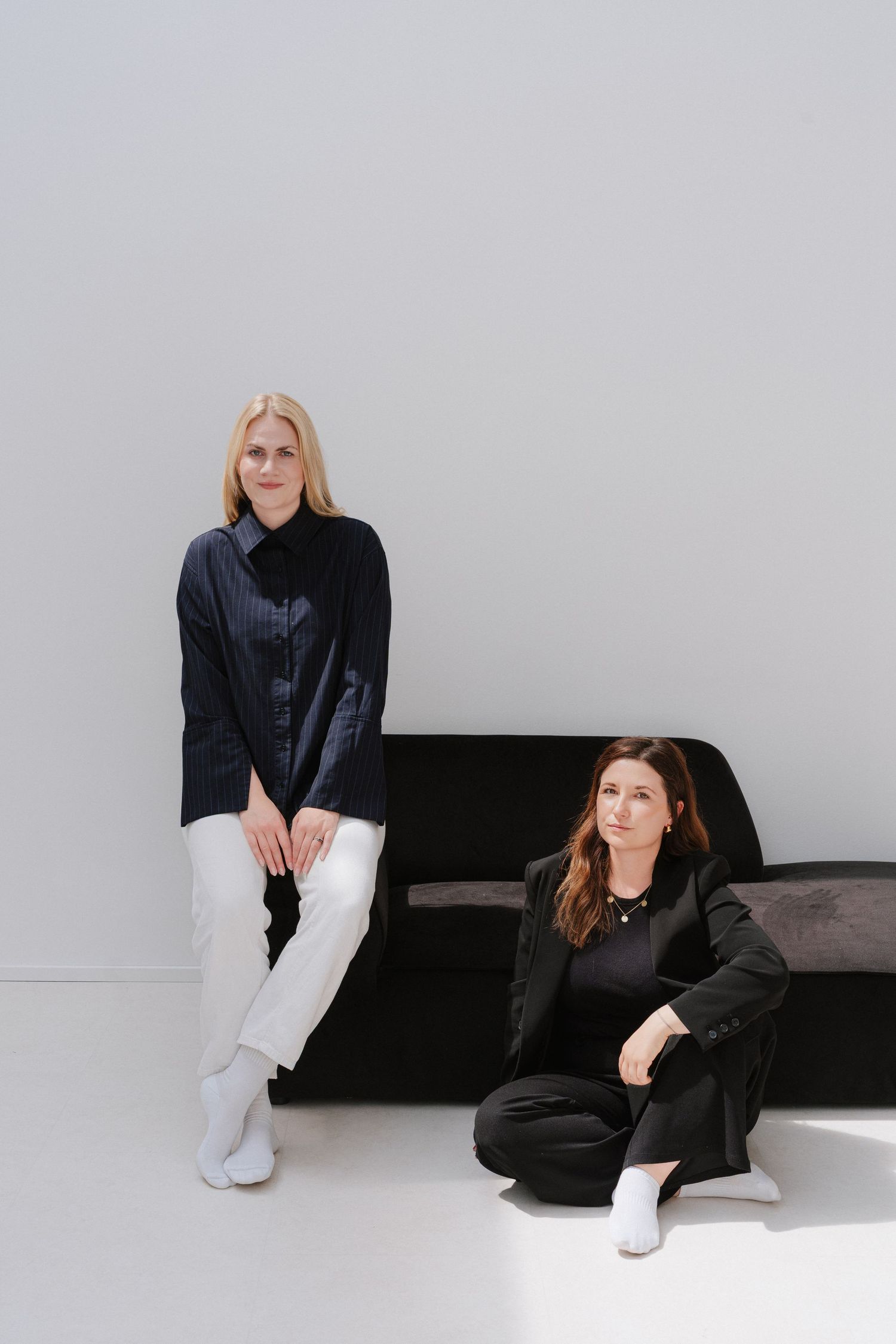 Two women in black and white outfits pose in a minimalist studio with a black sofa.