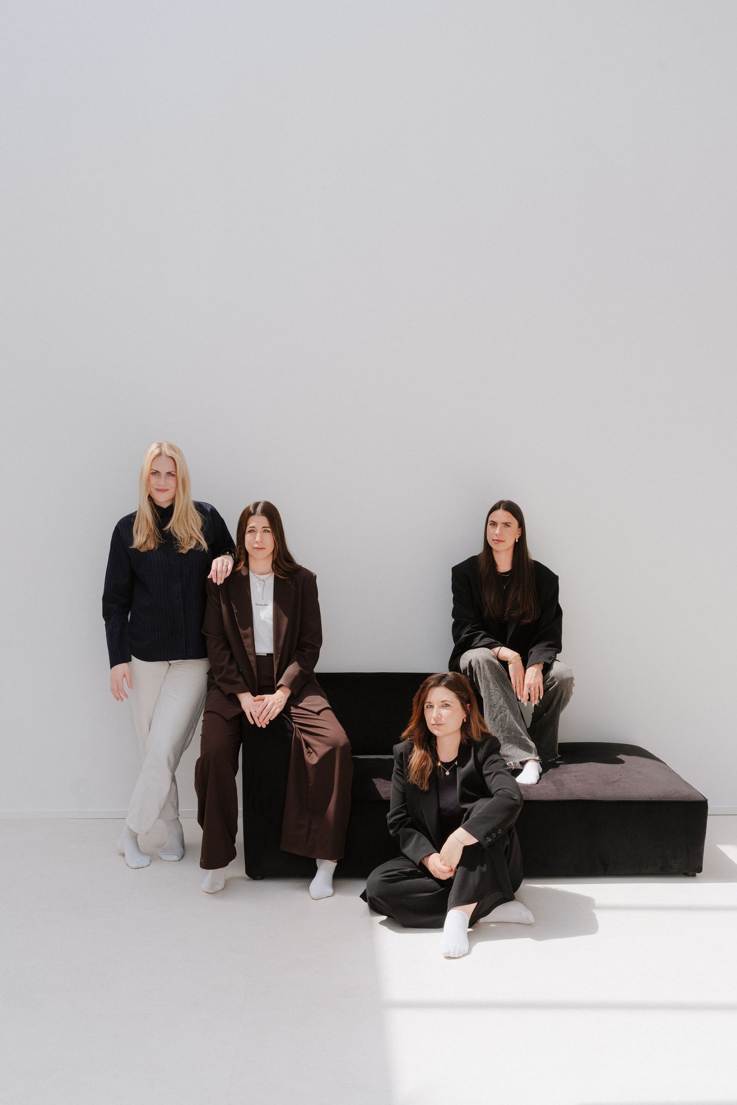 Four women in dark elegant outfits pose together in a minimalist studio with a black bench.