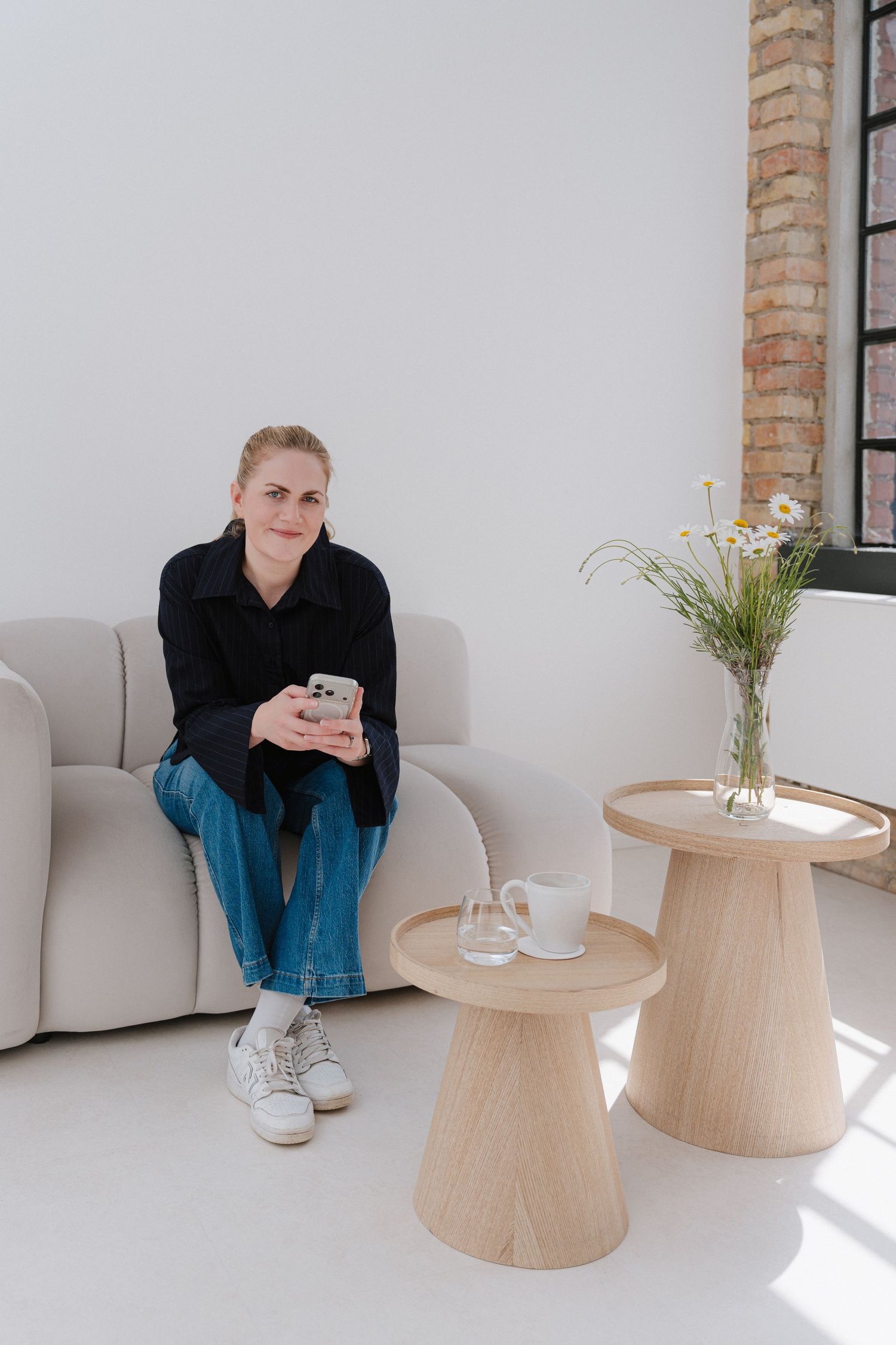 Woman sitting on a modern sofa holding a smartphone, with wooden side tables and a floral vase nearby.