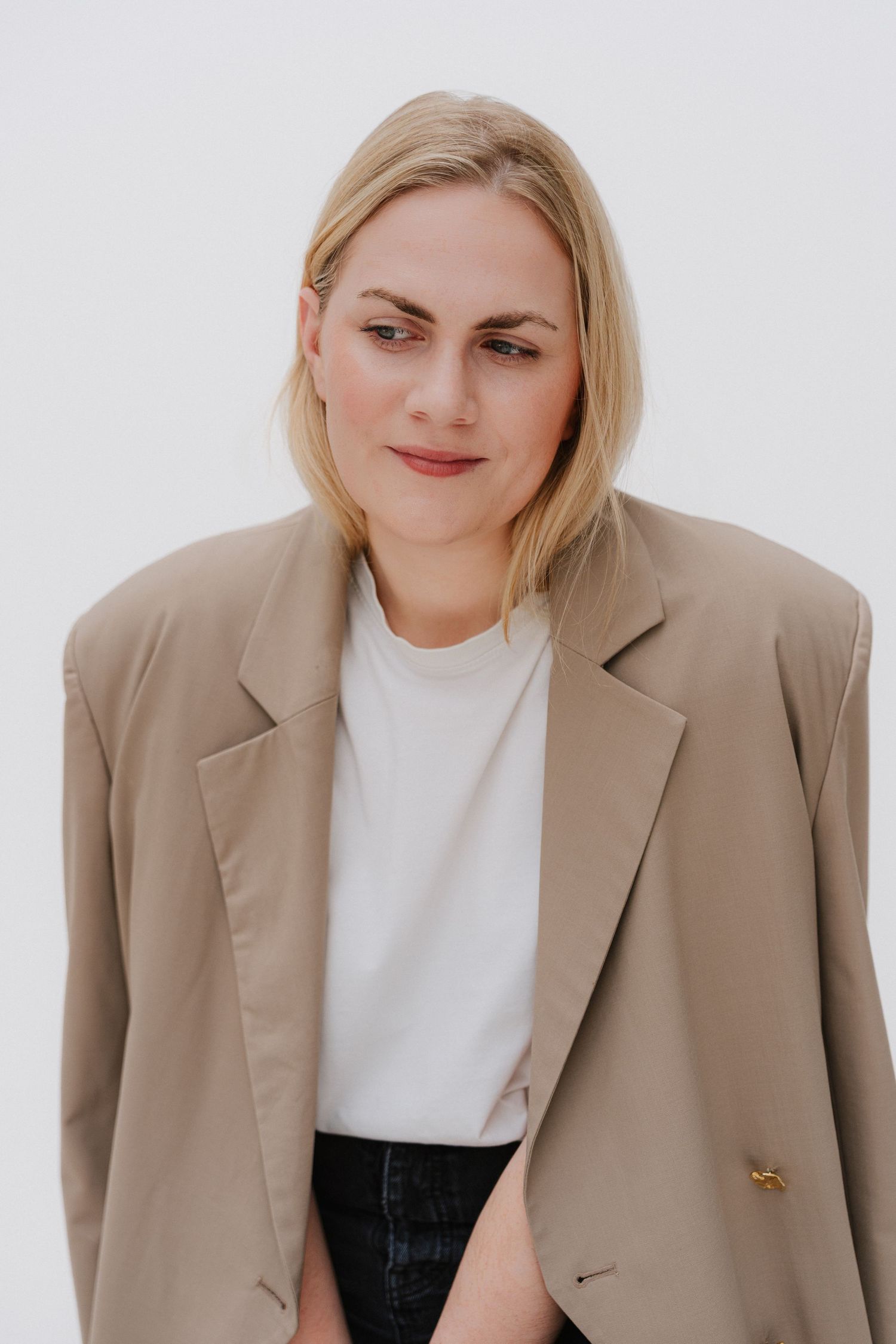 Blonde woman in oversized beige blazer and white t-shirt posing against a light background.