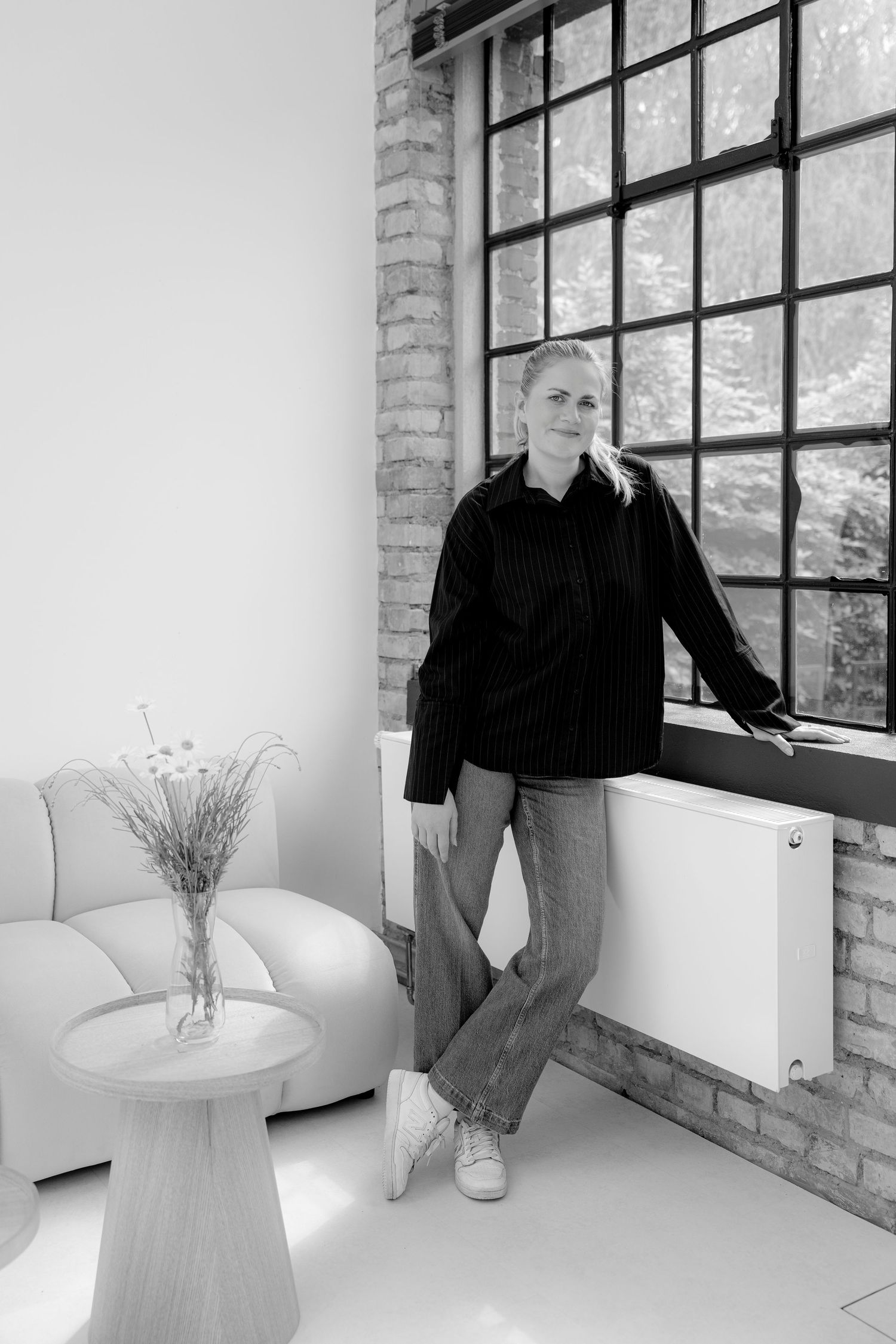 Black and white photo of a woman in jeans and oversized sweater leaning against a large industrial window in a modern loft.