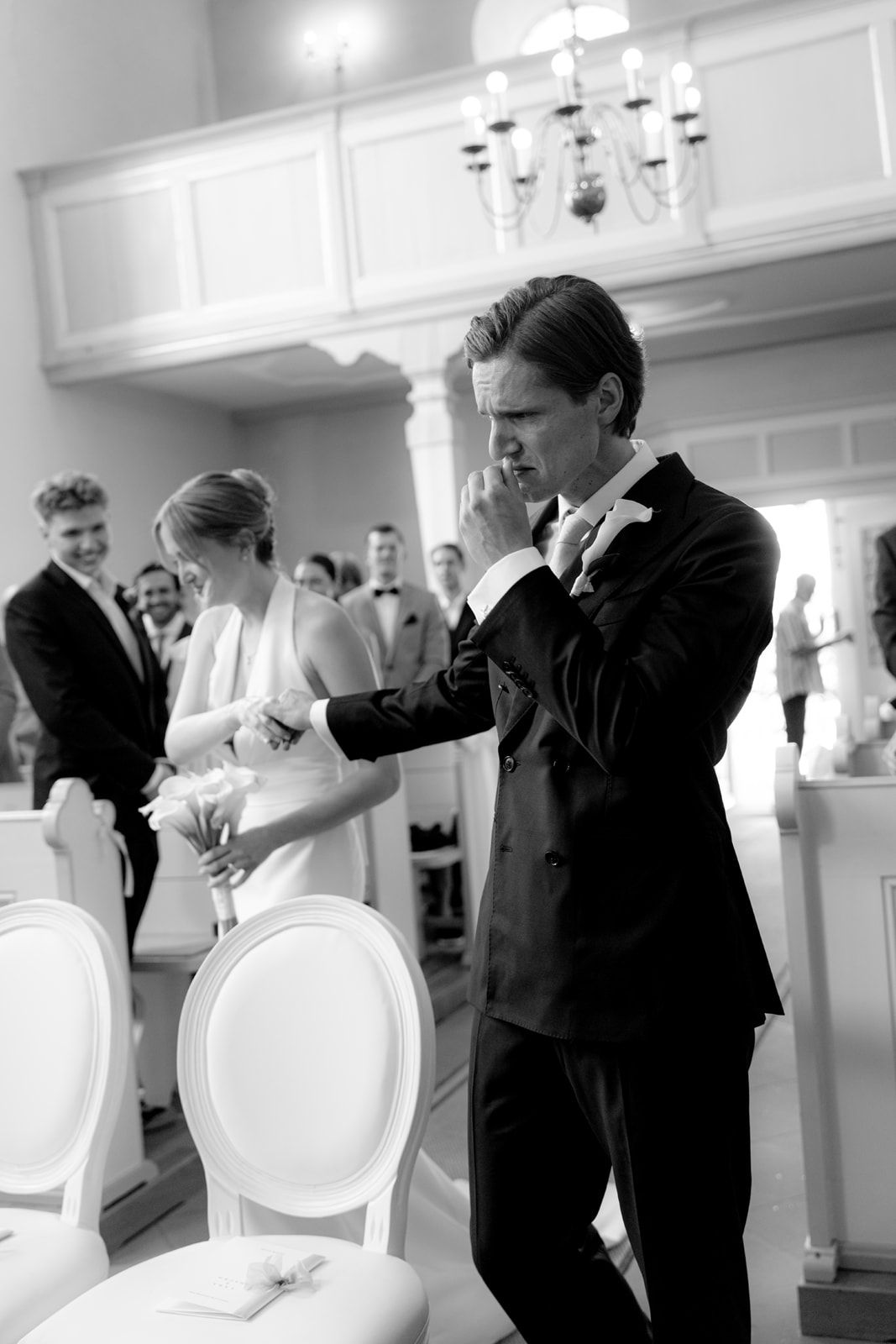 Emotional groom wiping tears during wedding ceremony in elegant white church, bride visible in background.