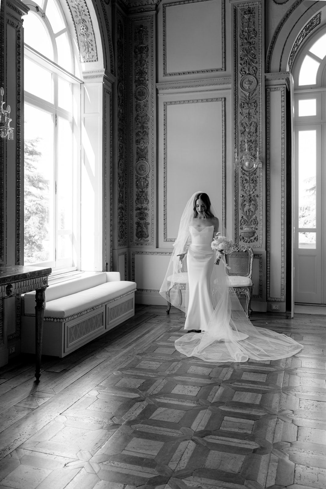Bride in white gown and veil stands in ornate black and white ballroom with arched windows and parquet floors.
