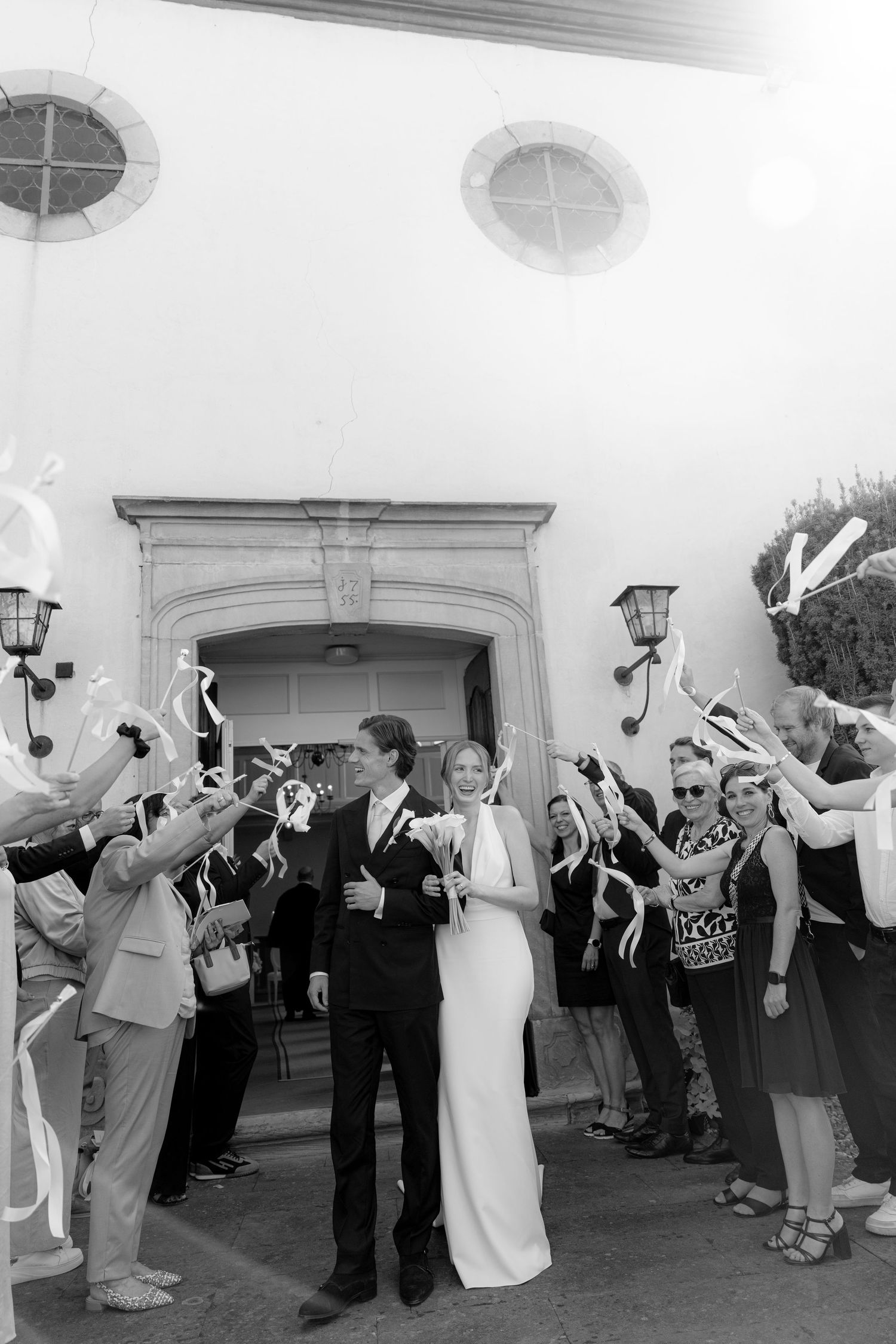 Black and white photo of a newlywed couple exiting a church while guests wave ribbon wands in celebration.