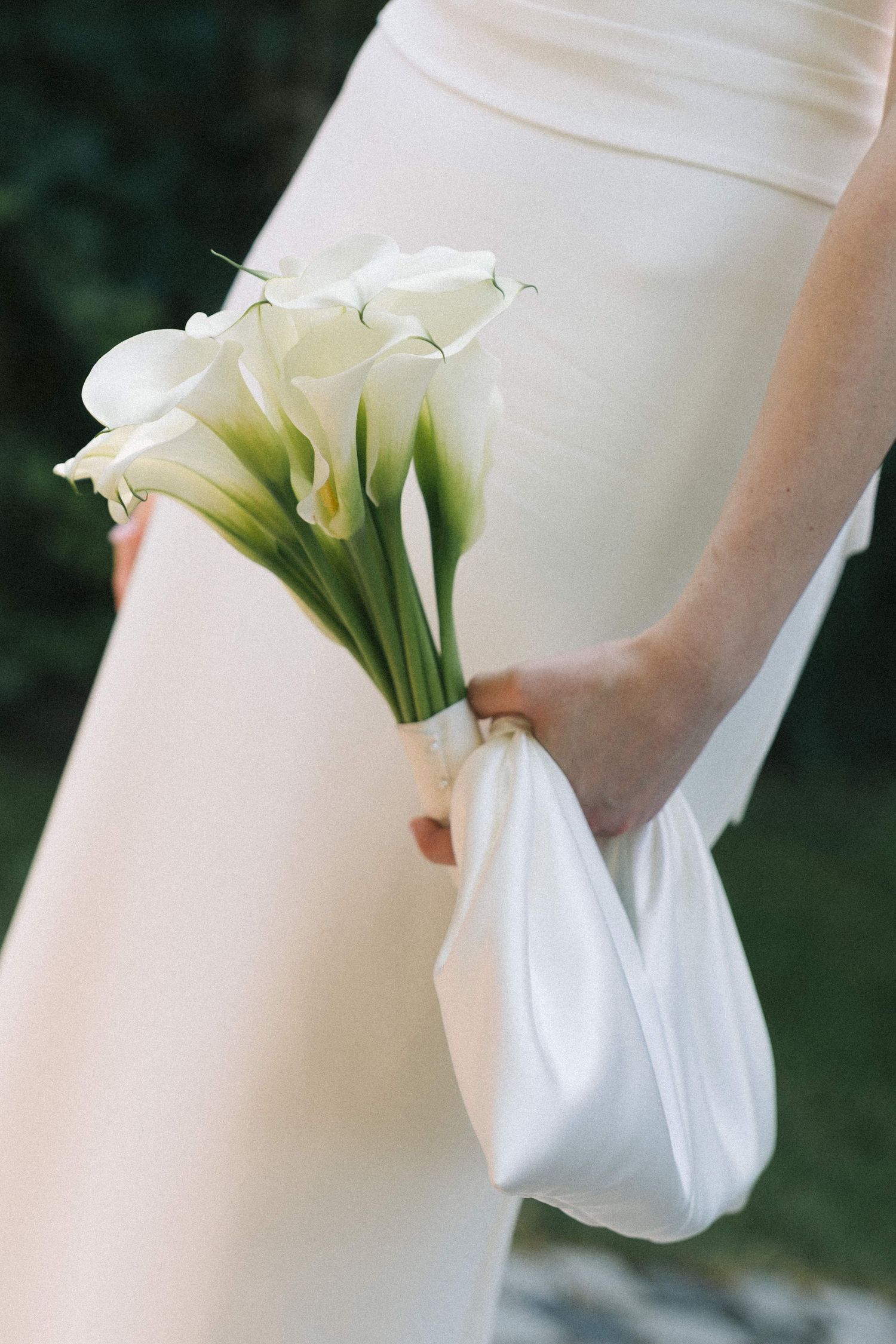 Bride holding elegant white calla lily bouquet wrapped in satin ribbon against her ivory wedding gown.