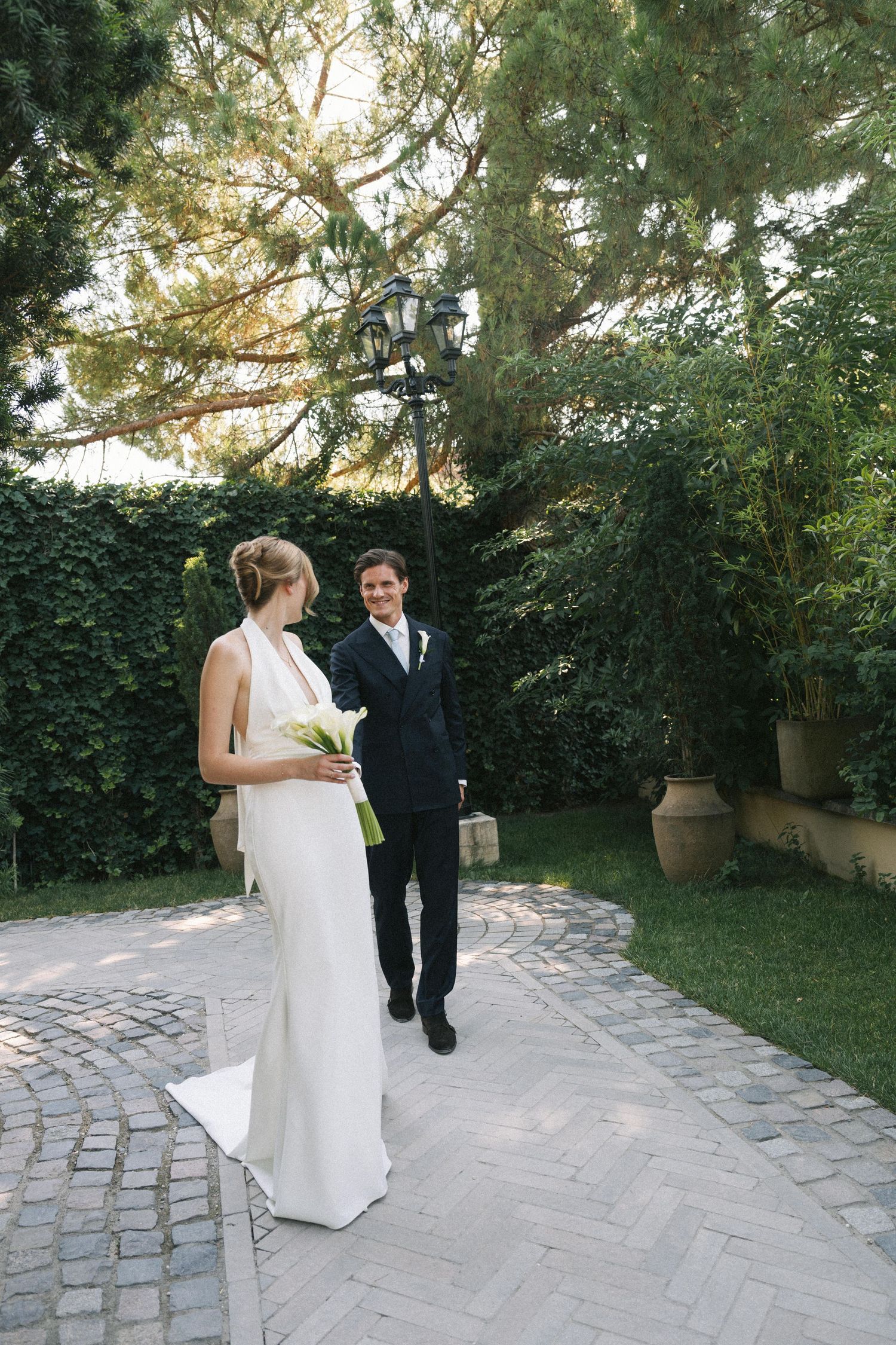 Bride in white gown holds bouquet as groom in dark suit smiles at her on a cobblestone garden path.