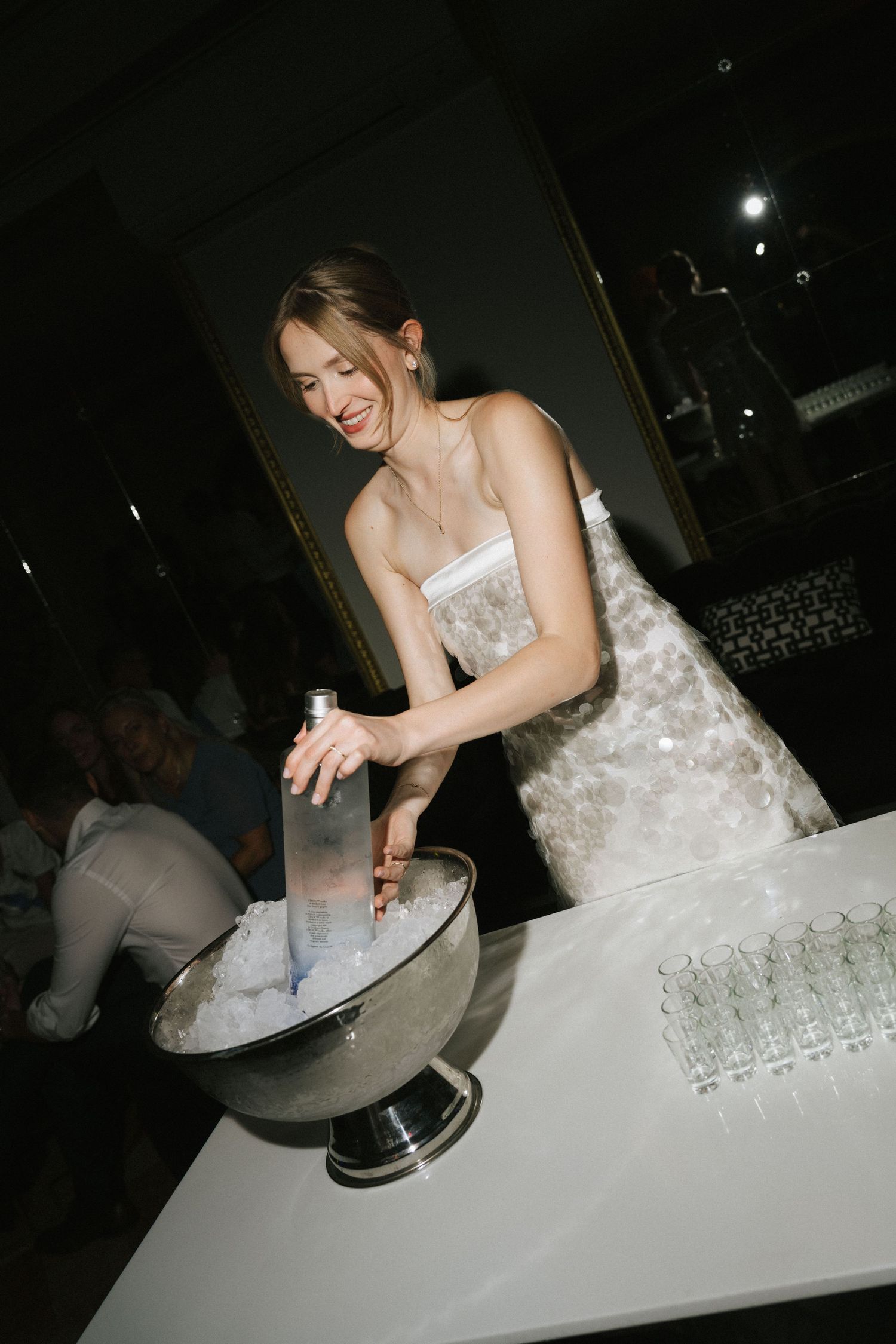Smiling bride in white gown placing bottle in ice bucket at wedding reception bar.