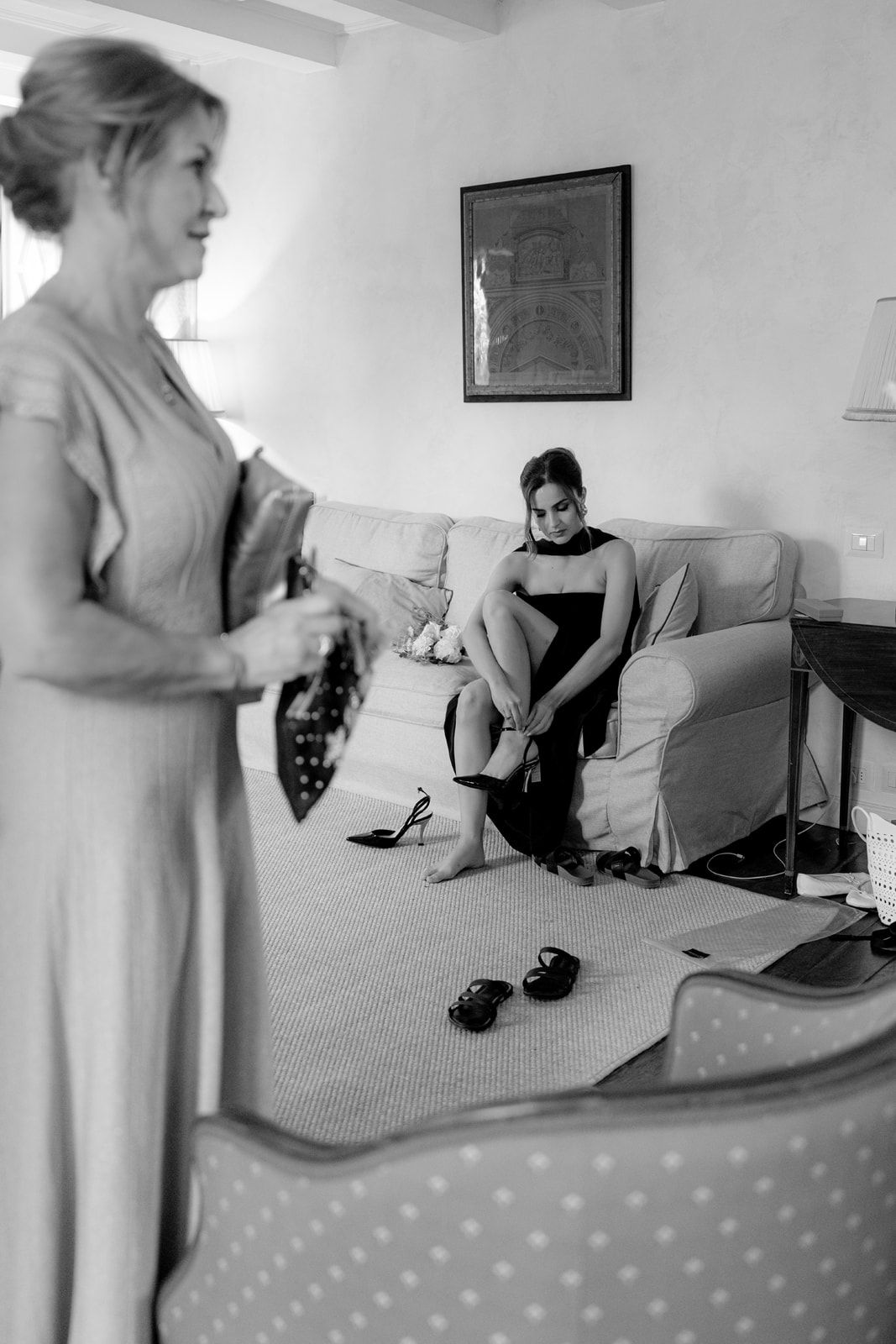 Black and white photo of two women getting ready, one adjusting heels on a sofa while another holds shoes in an elegant room.